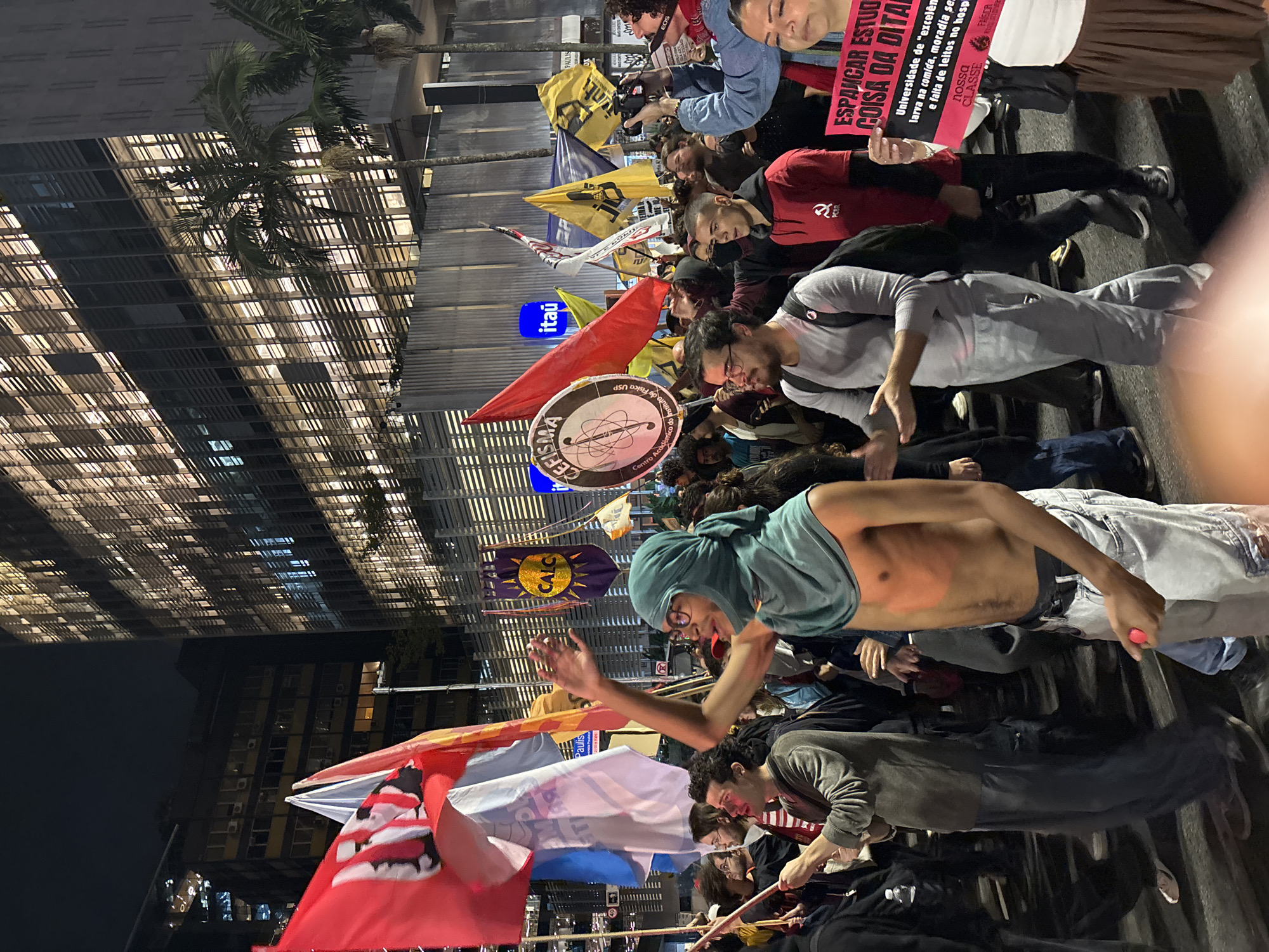 The image depicts a crowd of people gathered at night, seemingly engaged in a demonstration or protest. The scene is illuminated by streetlights and the lights from the surrounding buildings. The group is holding flags, banners, and signs with political messages, and includes an ITAU sign, and references to USP (University of São Paulo).
The demonstrators are a mix of ages. One male is shirtless and wearing glasses. Some hold flags.
The backdrop shows a modern city skyline with high-rise buildings. The event appears to take place in a public area.