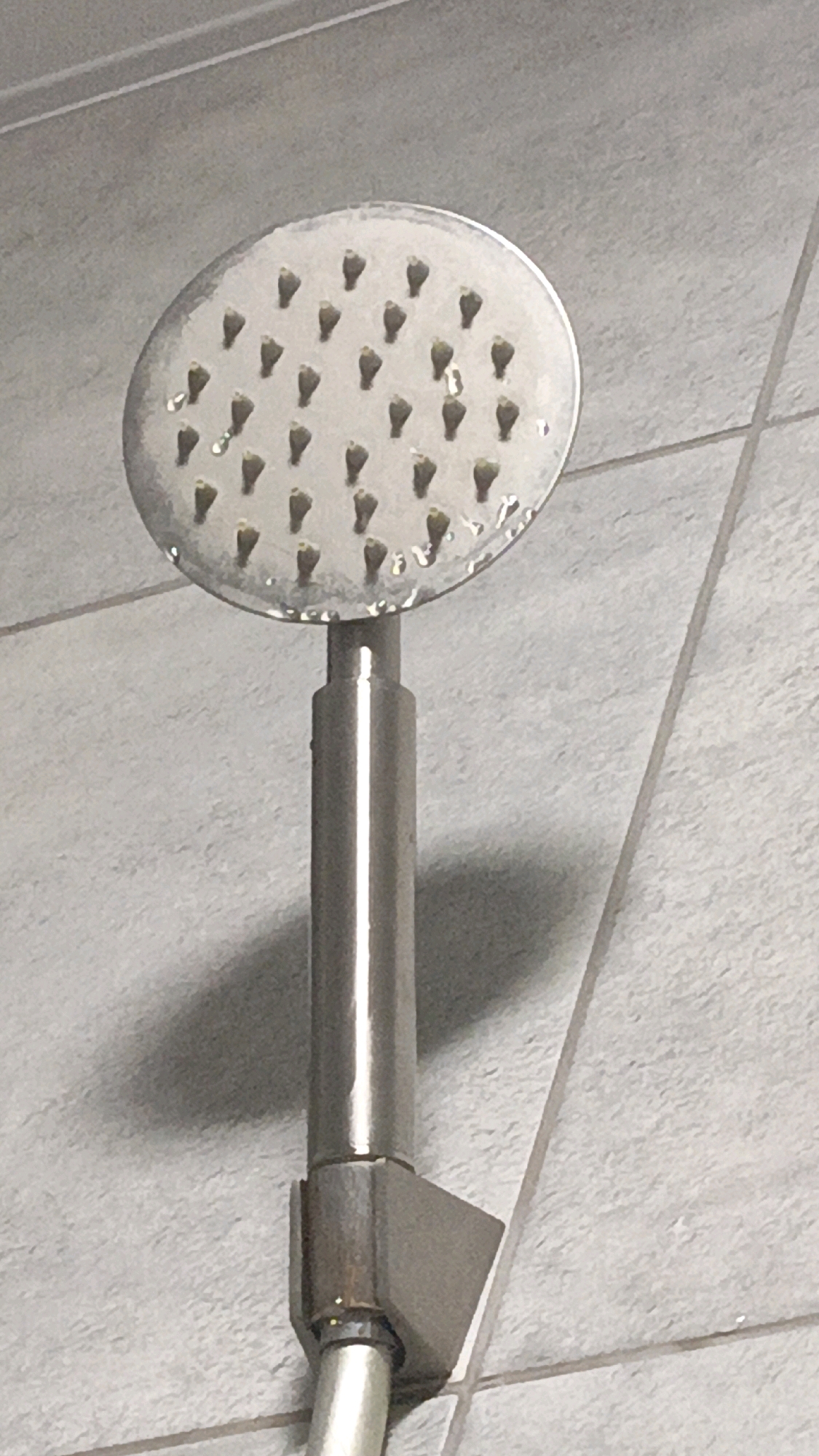 The image shows a close-up, slightly angled view of a chrome shower head with multiple small spray nozzles, some of which appear to have water droplets on them. The shower head is attached to a silver-toned metal arm, which then connects to a pipe. The background consists of textured, light gray tiled walls with visible grout lines. The lighting creates a shadow from the shower head and arm on the wall. The scene appears to be in a bathroom. There is no discernible information within the image to suggest a specific location in Homyel, Belarus.