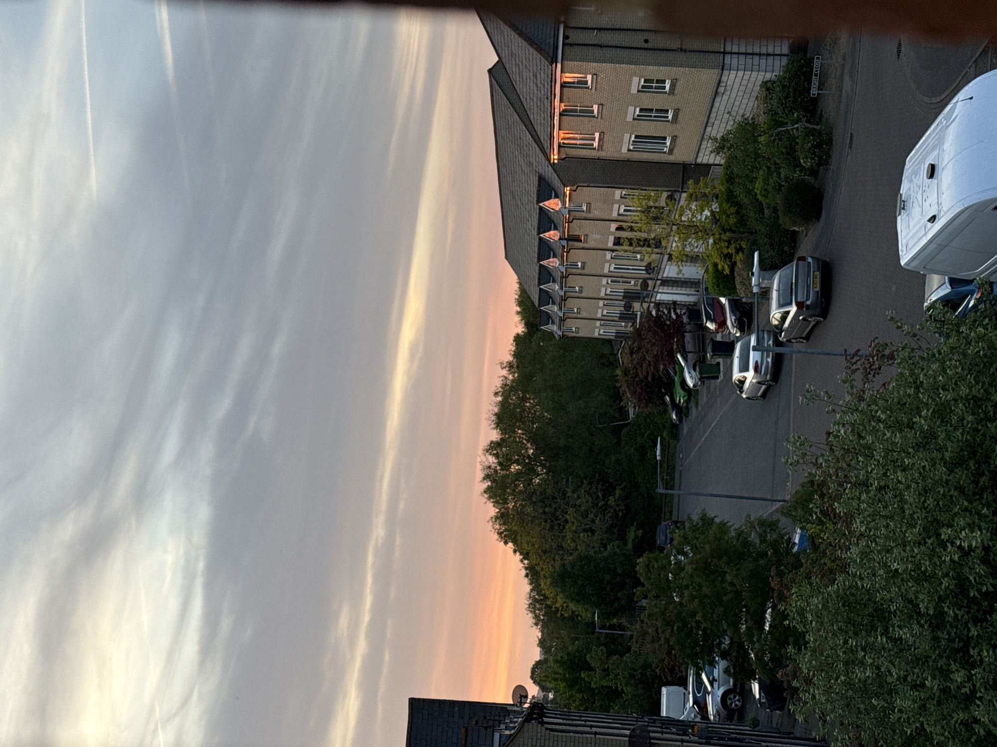 The image shows a street scene in Great Yarmouth, United Kingdom, at sunset. The sky is a mix of light gray and orange hues with wispy clouds. There are multi-story brick buildings with dark roofs and rows of windows. A street sign reads "BRIGHT CLOSE." Several cars and vans are parked along the roadside, nestled amongst green trees and bushes.