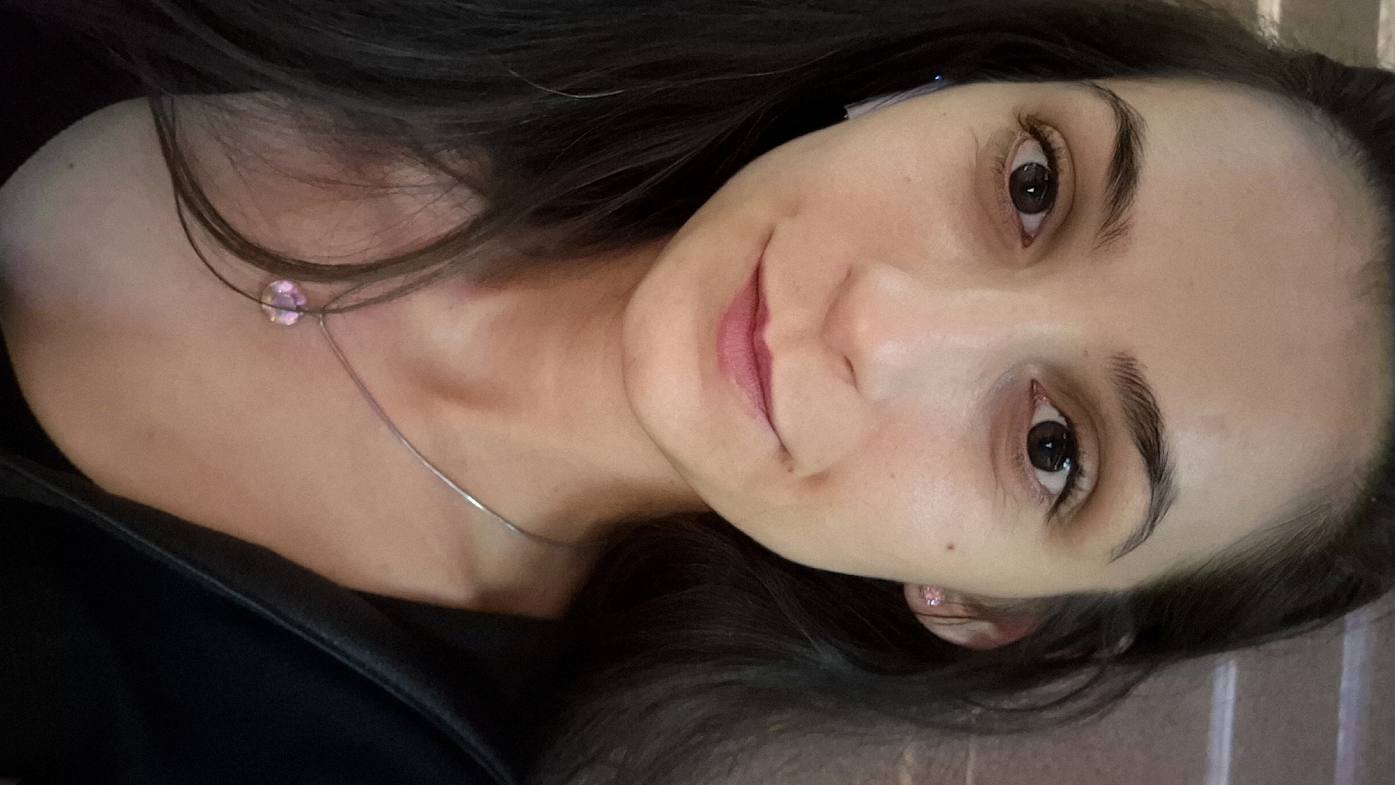 A close-up, head-and-shoulders selfie shows a young woman with dark hair, dark eyes, and a fair complexion. She is wearing a black top, a silver necklace with a light pink pendant, and small pink stud earrings. There is also a subtle hint of pink lipstick. The background is blurred and indistinct, providing no contextual information about the location. Without further information or visual cues, it is impossible to determine the specific location in Sofia, Bulgaria, or any other city.