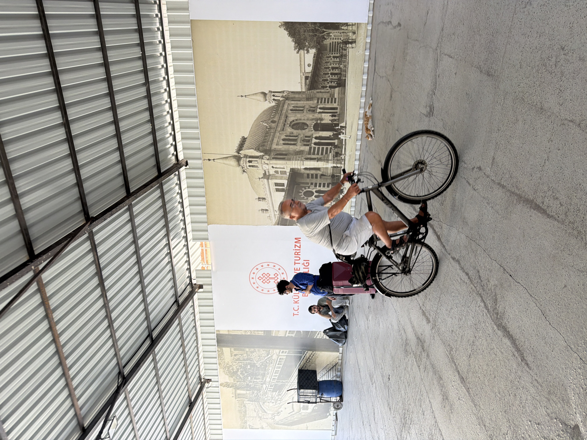 A middle-aged man with glasses and gray hair cycles across a concrete floor under a corrugated metal roof. He wears a gray t-shirt and white shorts. His bike is dark gray. Behind him, a large mural depicts a historical building. To the left, another man in a blue shirt stands near luggage, with two people sitting on the ground and bags nearby. A trolley with a blue barrel and crates is also visible. A ginger and white cat lies on the ground in front of the mural. The scene suggests a public space, possibly a transportation hub or exhibition area, in Istanbul, Turkey.