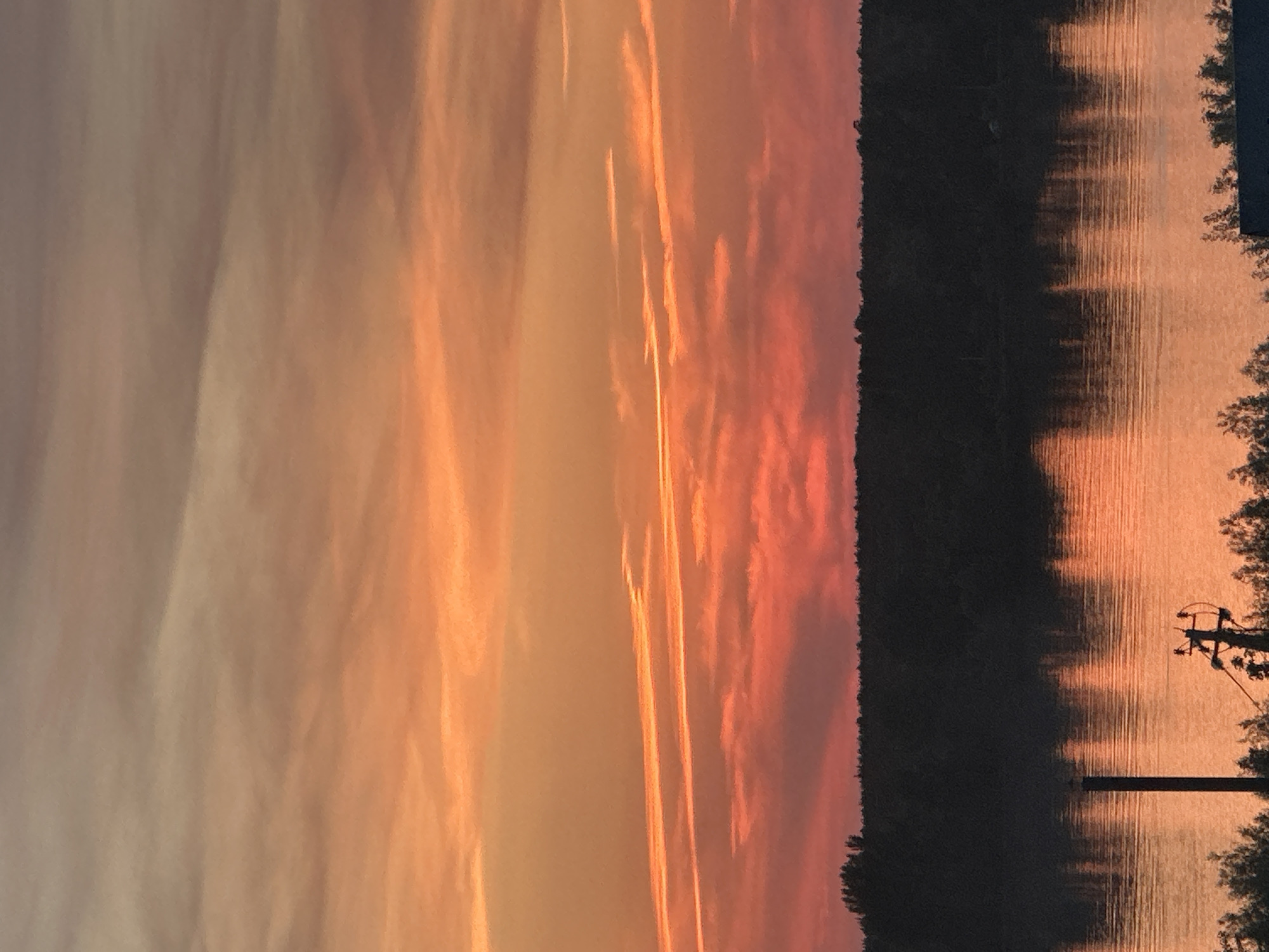 The image depicts a sunset over a body of water, likely a river or lake. The sky is filled with streaks of orange and pink hues, possibly with contrails. A dark treeline is visible on the right. The water surface reflects the sunset colors, creating vertical patterns. A utility pole with wires is seen in the right foreground. The image is rotated. The location might be Chaikovsky, Russia, but there's nothing in the photo to confirm it.
