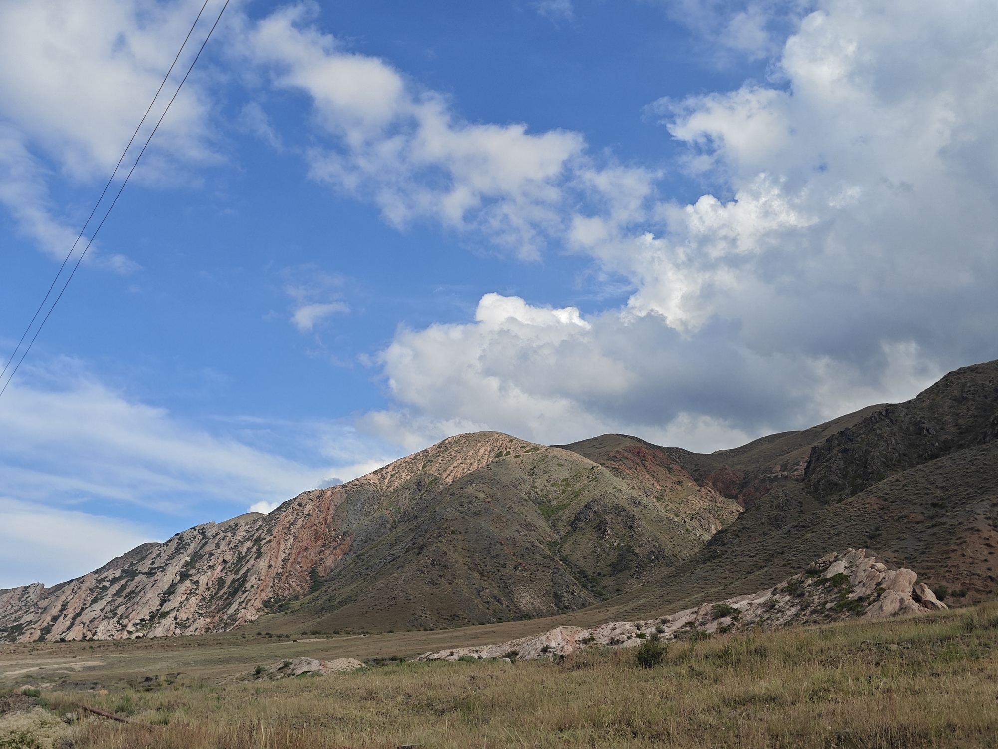 The image depicts a scenic view of rolling hills and mountains under a bright blue sky with scattered white clouds. The landscape is predominantly arid, with dry grass covering the foreground and lower slopes of the hills. The mountains themselves exhibit varied geological formations, with exposed rock faces showing bands of red and lighter-colored stone, suggesting stratification. In the far distance, a few dark specks, possibly vehicles or animals, are visible on a faint road. Overhead, a power line stretches diagonally across the upper left portion of the frame. The scene is natural and undeveloped, characteristic of a rural or mountainous region. No people are visible in the image. This vista is consistent with the landscape found near Kadji Say, Kyrgyzstan, a settlement known for its proximity to mountainous terrain and natural beauty.