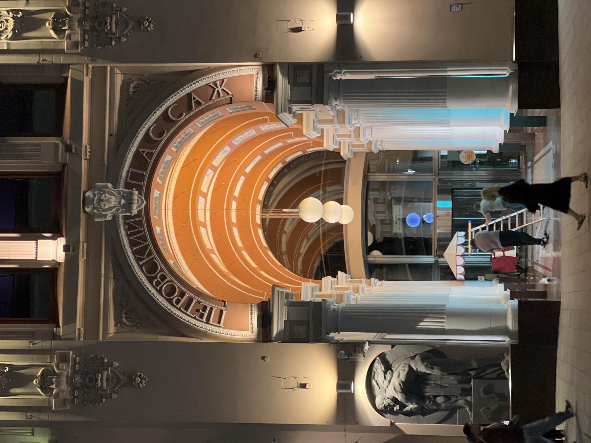 The image depicts the illuminated main entrance of Petrovsky Passage, a historic shopping arcade located in Moscow, Russia, at night. The central feature is a large, ornate archway with "ПЕТРОВСКИЙ ПАССАЖ" inscribed in Cyrillic letters above it. The arch's interior is characterized by a series of prominent orange-colored, ribbed arches, creating a vaulted tunnel effect.
Flanking the entrance are elaborate architectural details, including relief sculptures and decorative elements. To the left of the main arch, a large, dark grey bas-relief sculpture of two muscular male figures in a dynamic pose is mounted on the facade. Above the archway, intricate carvings of stylized lion heads, human-like faces, and scrolling foliage are visible. Inside the archway, several white, fluted columns line the path, with a long, vertical stack of three white spherical pendant lights hanging centrally.
The scene is lit by various fixtures. Wall-mounted cylindrical lights cast warm glows on either side of the facade, and an electric blue light emanates from a vertical strip on the rightmost visible column. Further inside the passage, through glass doors, two glowing blue spherical objects or lights are discernible.
Four individuals are present. In the left foreground, a person wearing dark clothing and blue jeans walks from left to right. In the center-right foreground, a person in a dark dress walks from left to right. Inside the passage, near a small red and white striped cart, a person in a light shirt and dark pants stands on a silver step ladder, facing away from the viewer. Another person in a light shirt is partially visible further back, near the glass doors, also appearing to be engaged in an activity related to the ladder or cart. The ground in the foreground is paved.