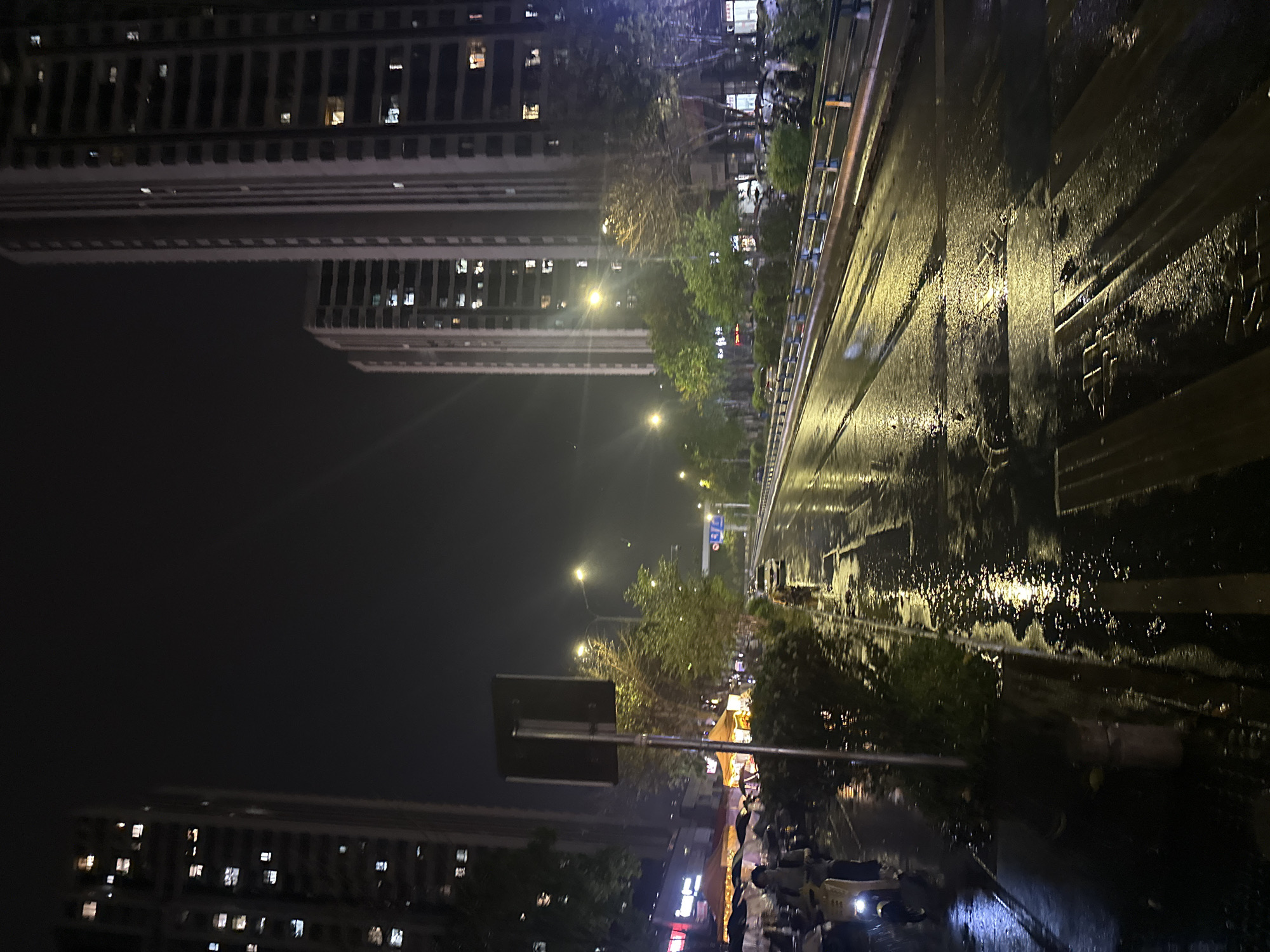 The image shows a nighttime street scene in Chongqing High-tech Zone, China, likely after a rain. The road is wet, with light reflecting off the surface, indicating water. On the right side of the frame is a street, probably a part of the main road. Buildings are visible on both sides. The buildings on the left are tall apartment complexes with multiple windows lit up. On the right, lights suggest shops or smaller buildings. There are several street lights along the road. In the foreground, there are parked vehicles or scooters. The general atmosphere is urban and damp. A traffic sign is visible. Overall, the scene suggests a quiet evening in a built-up area of Chongqing.