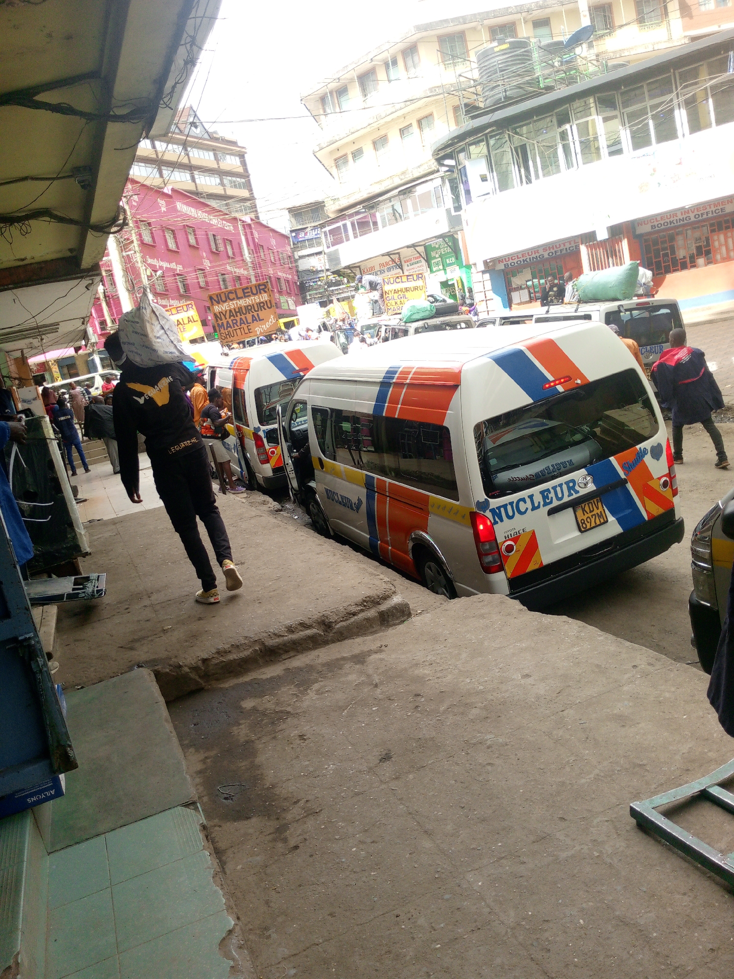 The image depicts a street scene in Nairobi, Kenya. Several white Toyota Hiace vans, marked with blue, orange, and red stripes, are parked along the side of the road, labeled with "NUCLEUR" and "Shuttle," indicating their use as public transportation vehicles. One van is identifiable by license plate "KDV 897M."
People are scattered around the vehicles and sidewalks. One individual is carrying a large bag over their shoulder, wearing a black jacket and pants with the text "LEGUATIZHE" on the leg. Other people are near the vans, some appearing to be getting in or out.
The street is cluttered with various objects, including what seems to be a metal frame lying on the ground. The buildings in the background are a mix of colors and styles, some with advertising for "NUCLEUR" investments and shuttles to locations like Nyahururu and Maralal. There are also signs for "KAIRI PARC" and "MOPESA" near a "NUCLEUR BOOKING OFFICE." The scene appears busy and indicative of a transport hub within a city environment.