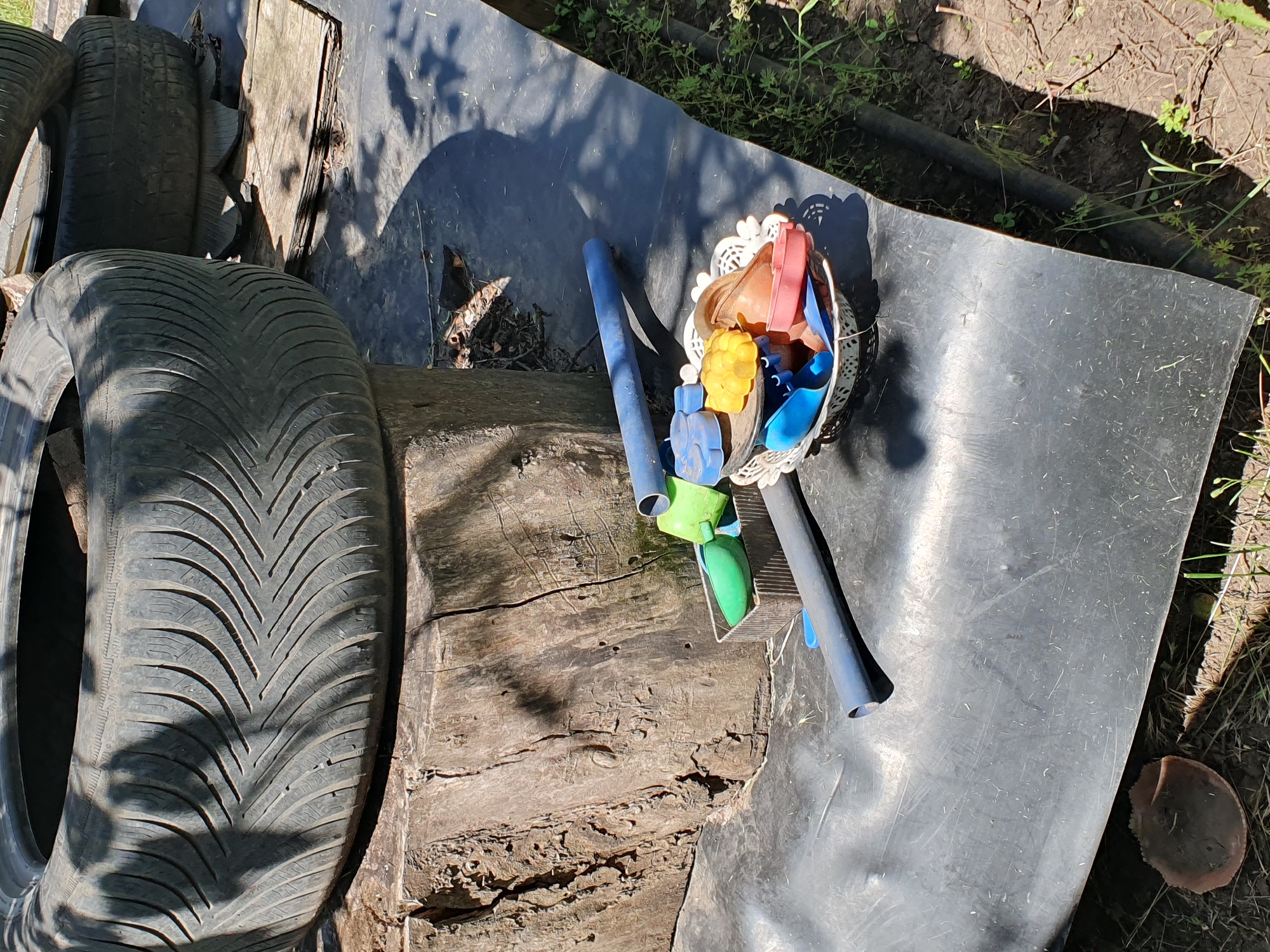 In Novoivanivka, Ukraine, a close-up, eye-level outdoor shot shows a collection of repurposed tires and gardening tools on a black plastic tarp. The foreground features a light-colored, lacy basket filled with small, colorful plastic scoops and molds in shades of blue, yellow, and red. Beside the basket, two dark blue plastic pipes lie horizontally, one in front of the other. To the left of the pipes, a small, rectangular metal container holds a bright green plastic scoop and a smaller blue scoop. A large, weathered wooden stump serves as a base for a portion of a black tire, which is partially obscured by shadows cast by unseen foliage. The tire has a distinct tread pattern. In the background, another tire rests on wooden planks. The ground surrounding the tarp is dry earth with sparse green vegetation and some scattered debris, including a dark, mushroom-like object near the bottom left corner of the frame. The scene appears to be a DIY garden or play area, possibly in a rural or suburban setting.