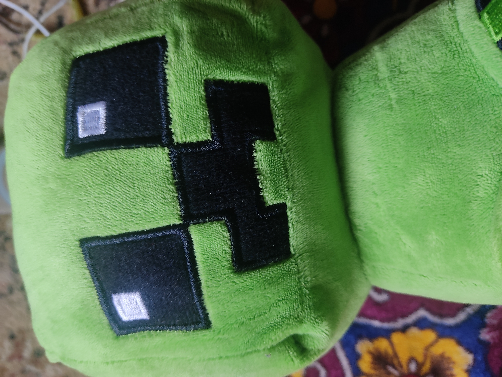 This media file shows a close-up of a stuffed toy that resembles a Creeper from the video game Minecraft. The toy is primarily a vibrant green color and has the characteristic Creeper face stitched onto it, consisting of black square pixels for the eyes and mouth. Within each eye, there is a smaller, lighter square patch, likely intended to represent a highlight or a different texture.
The stuffed toy is soft and appears to be made of plush material, with a visible fuzzy texture. It is positioned next to another similar green stuffed toy, only a portion of which is visible. The background is blurred and appears to be a surface with a colorful, patterned fabric, possibly a rug or blanket, featuring floral motifs in shades of yellow, blue, and pink. The lighting suggests an indoor setting, and the overall mood is playful and casual. There is no visible text in the image.