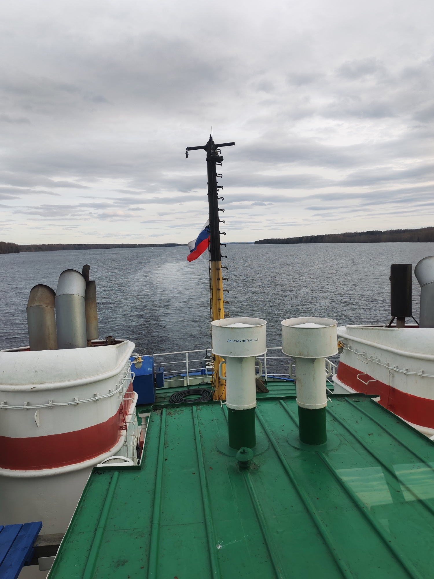 The image depicts a view from the deck of a vessel on a body of water. In the background, land is visible, potentially indicating the shoreline. The deck is green and features various shipboard elements, including white lifeboats with red striping, cylindrical vents, and a mast with the Russian flag. The sky is overcast with grey clouds. The location is in the city of Yakunina Gora, Russia.