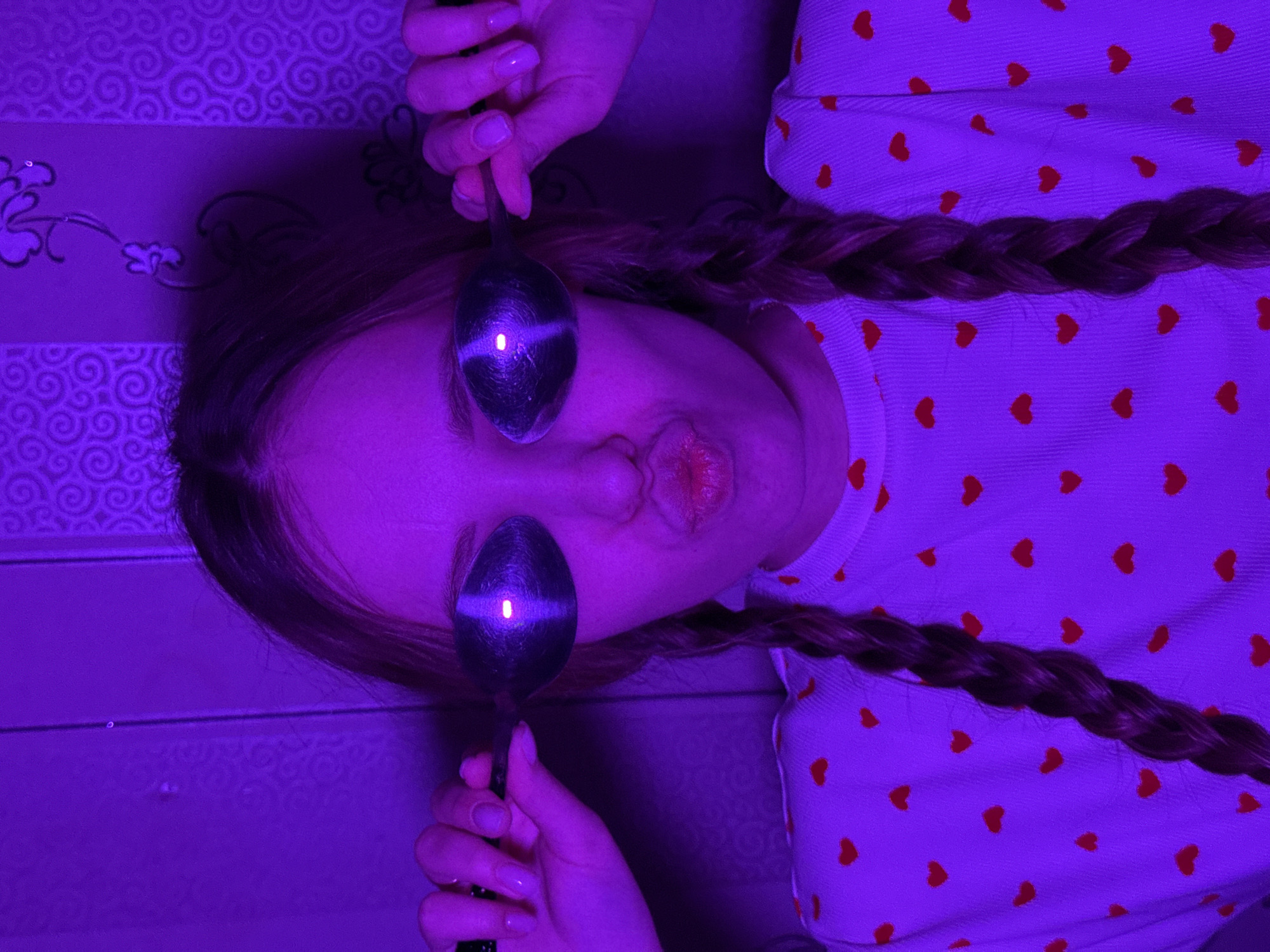 The image displays a close-up indoor portrait of a light-skinned individual, illuminated predominantly by purple light. The person, appearing female, is centered in the frame, facing forward with a pouting expression, lips pursed. Two metallic spoons are held by their handles, one over each eye, completely obscuring them. The bowls of both spoons reflect a distinct, bright vertical line of light, indicating a light source directly in front. The individual has long hair styled in two braids, which rest over her shoulders. She wears a light-colored garment, possibly a top or sweater, patterned with numerous small red heart shapes. Her hands are visible, with manicured fingernails showing a light-colored polish. The background consists of a wall covered with patterned wallpaper, featuring swirling floral motifs on the sides and a larger, distinct floral design centrally behind the subject's head. The specific location within the city of Chaikovsky, Russia, cannot be determined from the visual information provided.