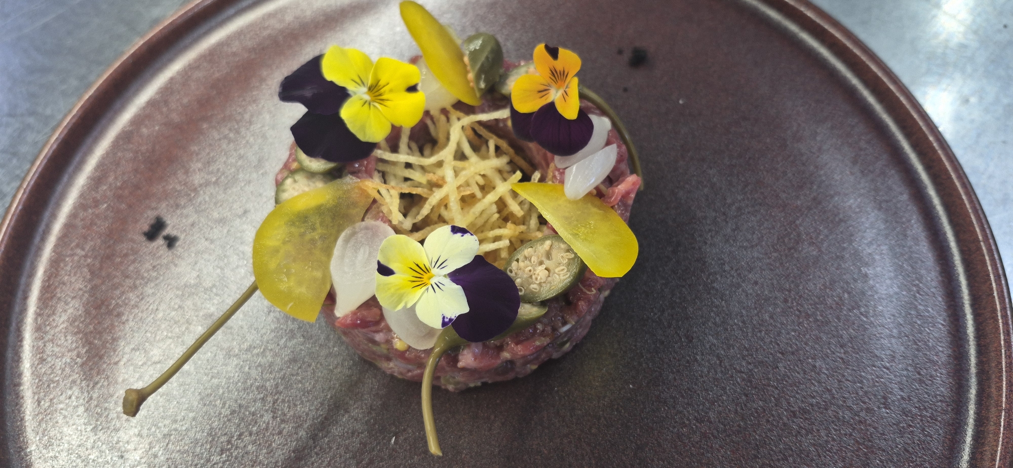A small serving of tartare is presented on a dark, textured plate. The tartare is topped with thinly sliced fried potatoes, pickled items that appear to be capers and possibly zucchini, and small yellow and purple edible flowers. A thin skewer emerges from the tartare, topped with a single yellow petal. The lighting is direct, creating some shadows and highlighting the textures of the food and plate. The background is a muted gray surface, possibly a countertop or table. The image is taken from a slightly elevated, top-down perspective. There are no people visible in the image. The location in Saint Petersburg, Russia, is not discernible from the visual information provided in the image.