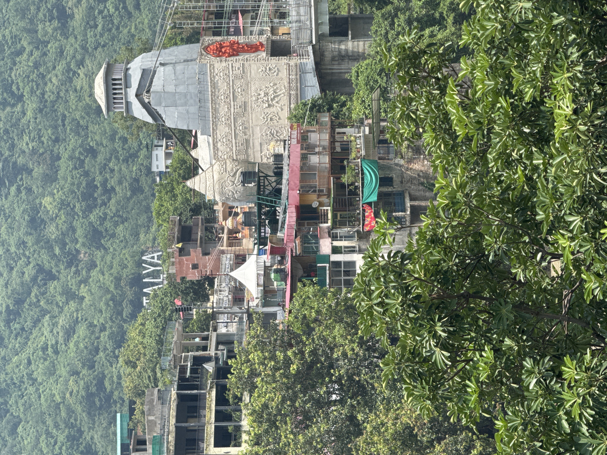 The image depicts a hillside urban landscape in Rishikesh, India, characterized by a dense, green, forested mountain in the background. In the mid-ground, a cluster of multi-story buildings with varied architectural styles is visible. Prominently featured is a large, gray, temple-like structure with intricate carvings and a towering finial. Adjacent to it are smaller buildings, some appearing to be shops or residences, with colorful awnings and balconies adorned with plants. A white tent is also visible among the buildings. In the foreground, lush green trees with broad leaves create a natural frame for the scene. The overall impression is one of a vibrant town nestled within a natural, hilly environment.