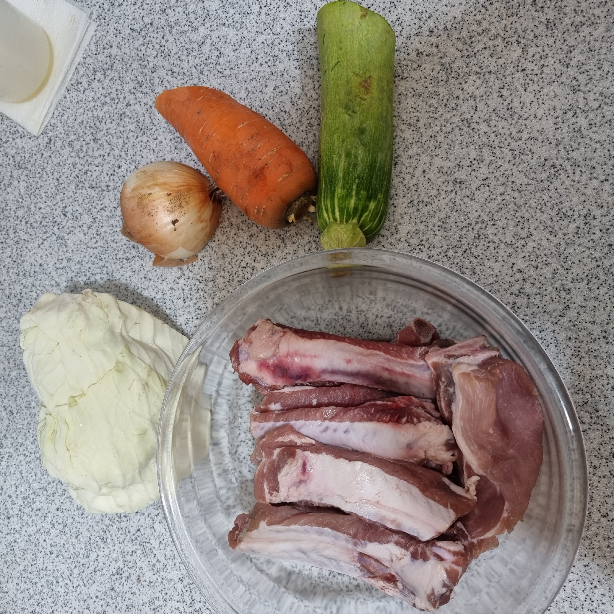 In Dunnae-myeon, South Korea, a flat, speckled grey countertop displays several raw food items arranged for preparation. In a clear glass bowl on the left are several racks of uncooked pork ribs. To the right of the bowl, a head of white cabbage sits at the top. Below the cabbage, a single yellow onion is visible. Further to the right and slightly below the onion, an orange carrot is positioned diagonally. At the bottom right, a green zucchini with visible striped markings lies horizontally. The overall scene suggests the preparation of a meal.
