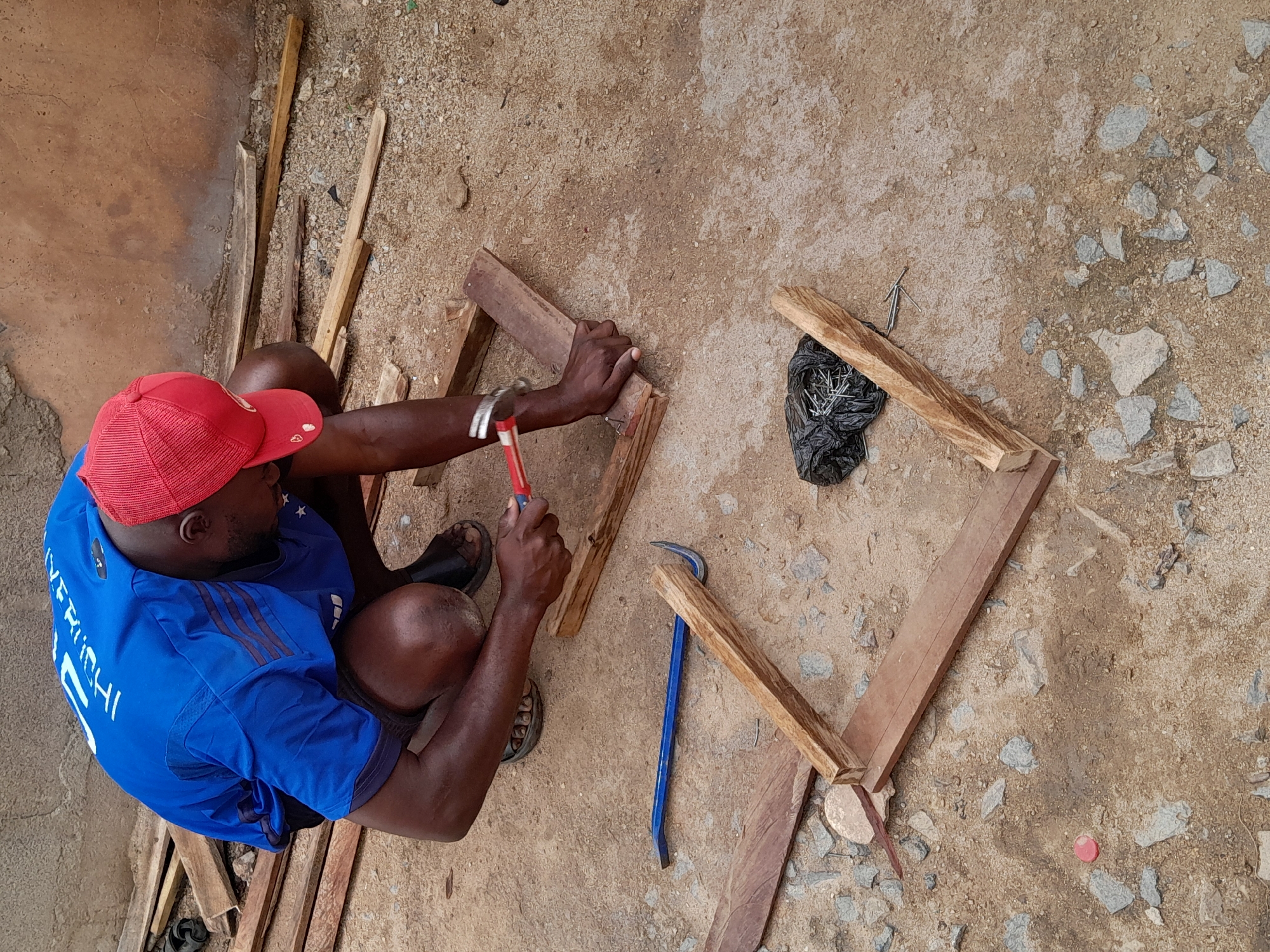 An overhead shot shows a dark-skinned man in Jos, Nigeria, squatting on a dirt and stone surface. He wears a red baseball cap, a blue soccer jersey with white lettering, and sandals. He is hammering together wooden pieces to form a rectangular frame. Scattered around him are additional wooden planks, a blue crowbar, a black plastic bag containing nails, and a red bottle cap. The background includes a textured wall.