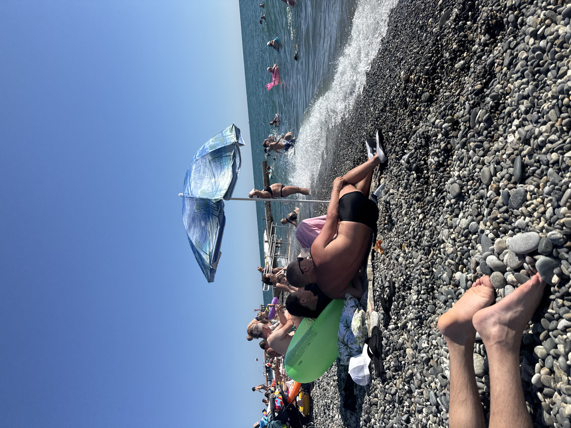 In Sochi, Russia, a pebble beach is bustling with people enjoying a sunny day. The scene is dominated by the azure expanse of the Black Sea, with waves gently lapping at the shore.
In the foreground, a couple is relaxing on the beach. A man, shirtless and wearing black swim trunks, lies on his back, with his head propped on a pink towel. Next to him, a woman with dark hair is leaning on her arm, looking towards him. A large, green inflatable raft lies nearby, and a white baseball cap and flip-flops are visible on the pebbles.
Behind them, a blue and white patterned beach umbrella offers shade to a woman in a black bikini who stands near the water's edge. Further out in the sea, numerous people are swimming and playing, some with inflatable rings and toys. A pink, fish-shaped float is particularly noticeable.
In the background, a wooden pier extends into the sea, with more people visible on it. The overall atmosphere is one of a lively and enjoyable summer day at the beach.