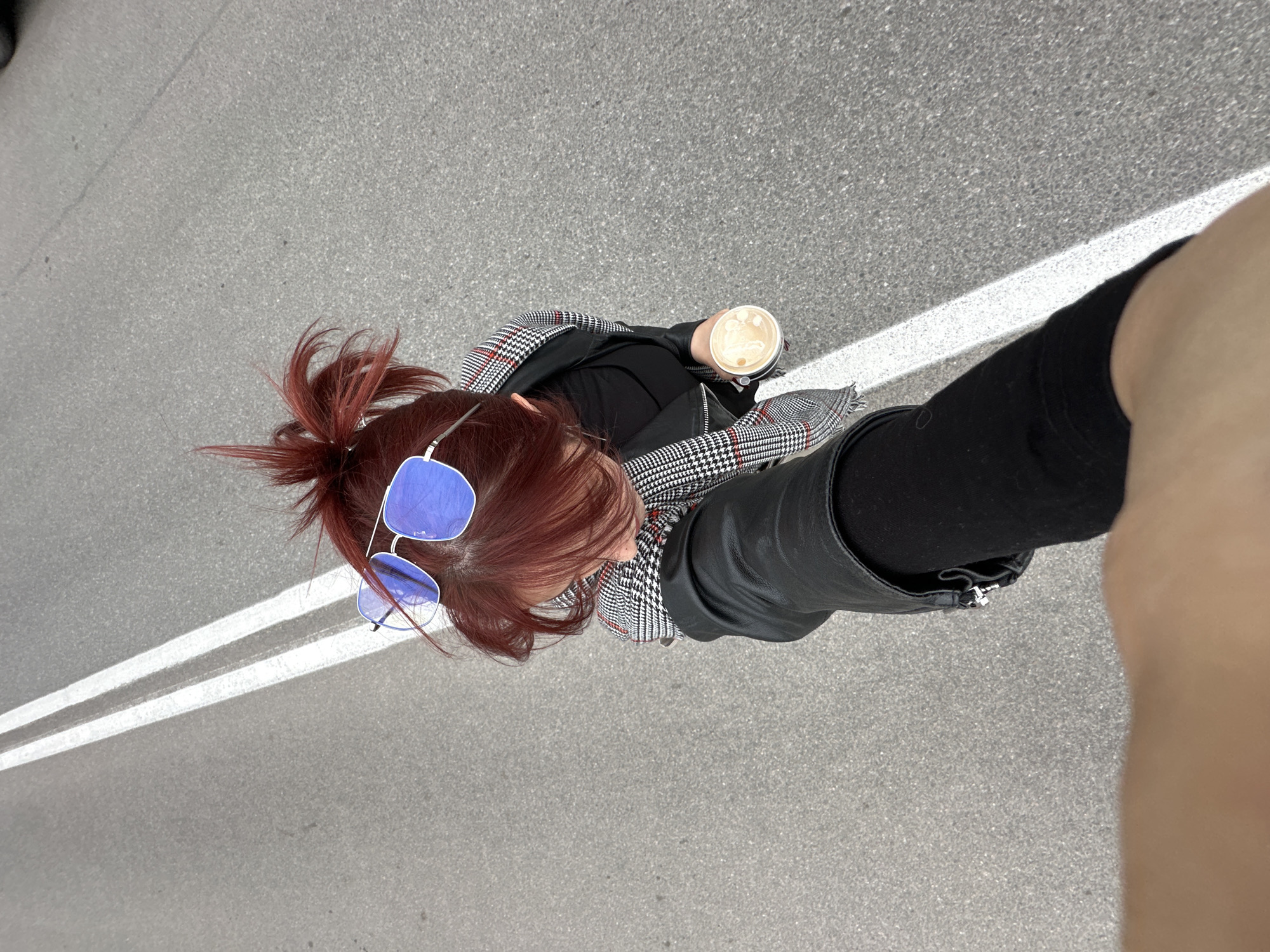 An overhead shot shows a person standing on a gray asphalt surface with two white parallel lines running diagonally across the image. The person has reddish-brown hair styled in a bun, and they are wearing blue sunglasses, a black top, and a black and white checkered blazer. They also have on a black leather boot on the left leg. The person is holding a cup, likely filled with coffee, in their right hand. The right leg is partially visible on the right side of the frame, showing a tan skin tone. The setting appears to be an urban environment, potentially a street in an unknown city.