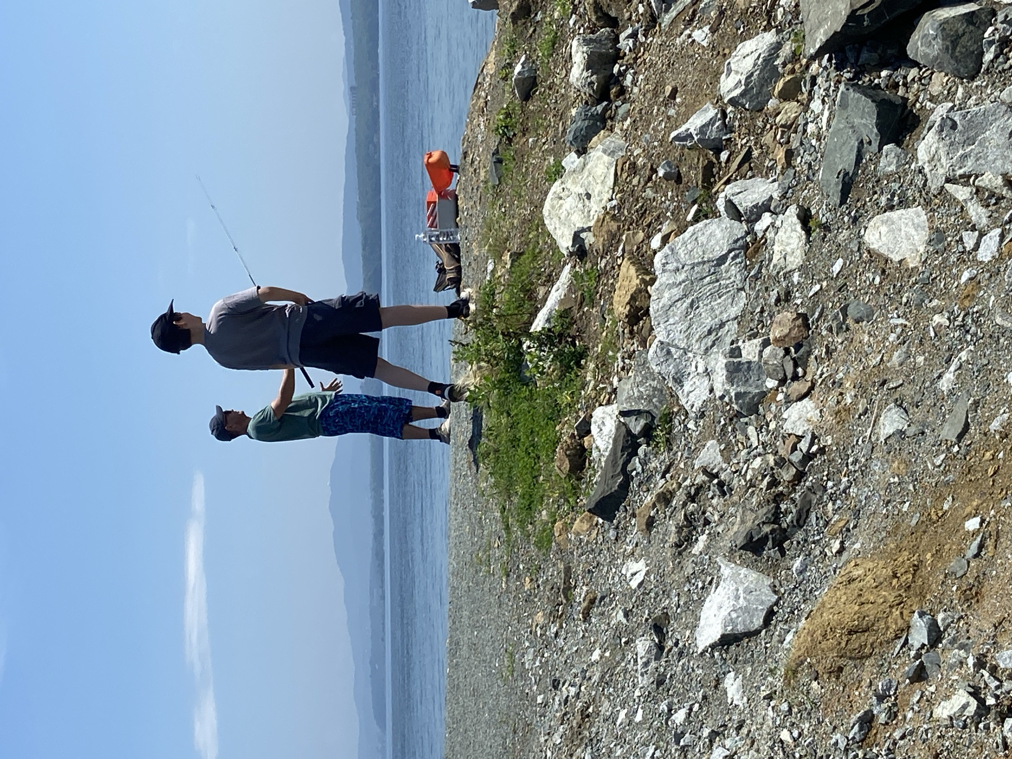 The image depicts two individuals, likely young men, fishing by a body of water. The person on the left wears a green t-shirt and blue patterned shorts, with a dark baseball cap and sunglasses. The person on the right wears a grey t-shirt, dark shorts, and a black baseball cap. Both are seen from behind, with the person on the right holding a fishing rod. A red kayak and a water bottle are placed on the rocky shore behind them. The environment is characterized by a rocky, pebbly shoreline leading down to calm blue water, with distant, hazy mountains on the horizon under a clear blue sky. The scene suggests an outdoor recreational activity.