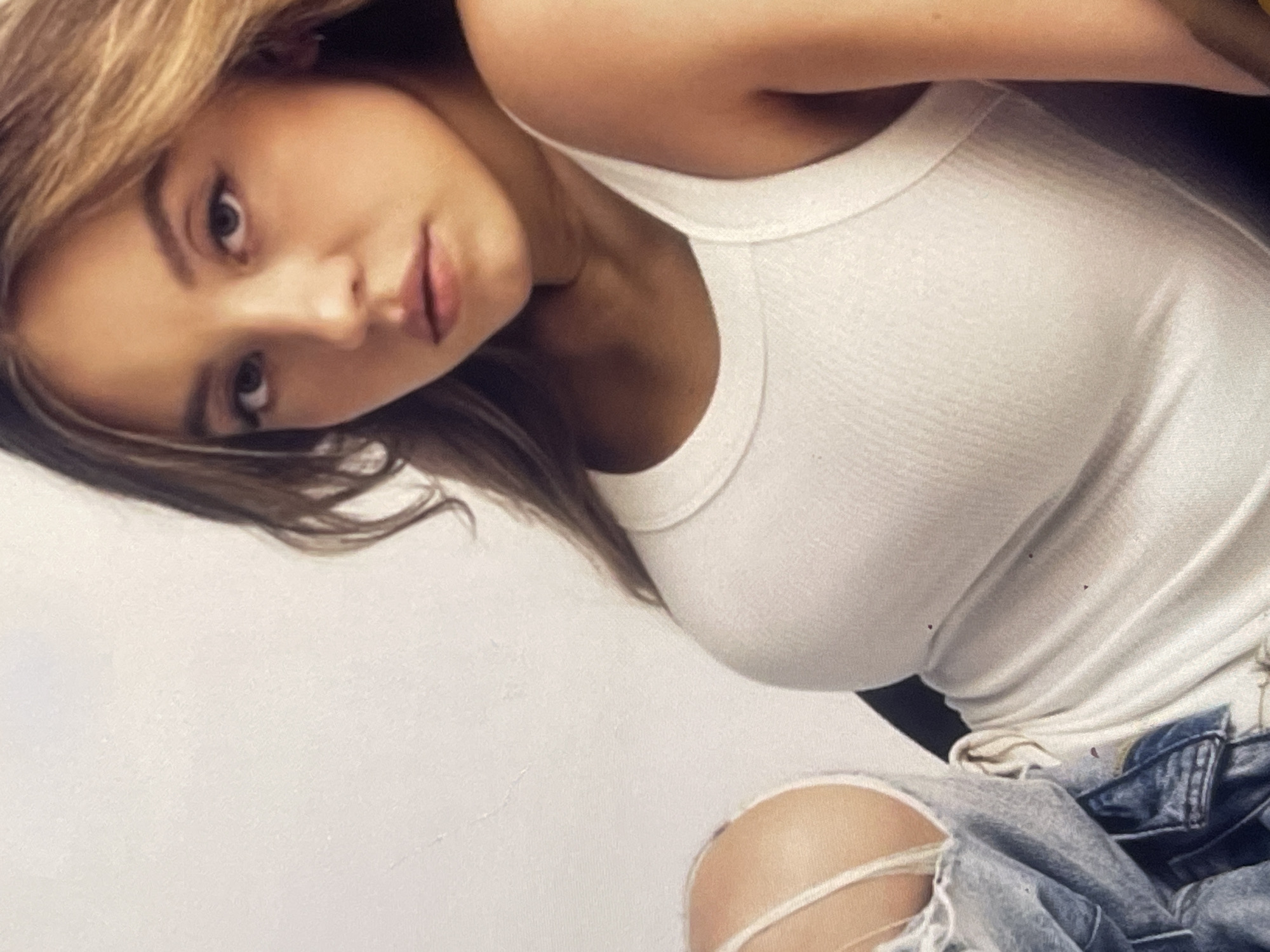 A young woman is captured in a close-up, slightly tilted portrait, possibly a selfie. She has light brown or blonde hair, styled to frame her face, and is looking directly towards the camera with a neutral, perhaps slightly alluring or pensive, expression. Her lips are slightly parted. She is wearing a white, ribbed tank top that reveals her shoulders and collarbone. Below her waist, torn blue jeans are visible, with a prominent rip over her left knee. The background is a plain, light-colored surface, likely a wall, suggesting an indoor setting. No specific location cues are visible within the image itself to confirm the provided context of Poltava, Ukraine. There is no visible text. The lighting appears soft and even, highlighting her facial features and the texture of her clothing.
