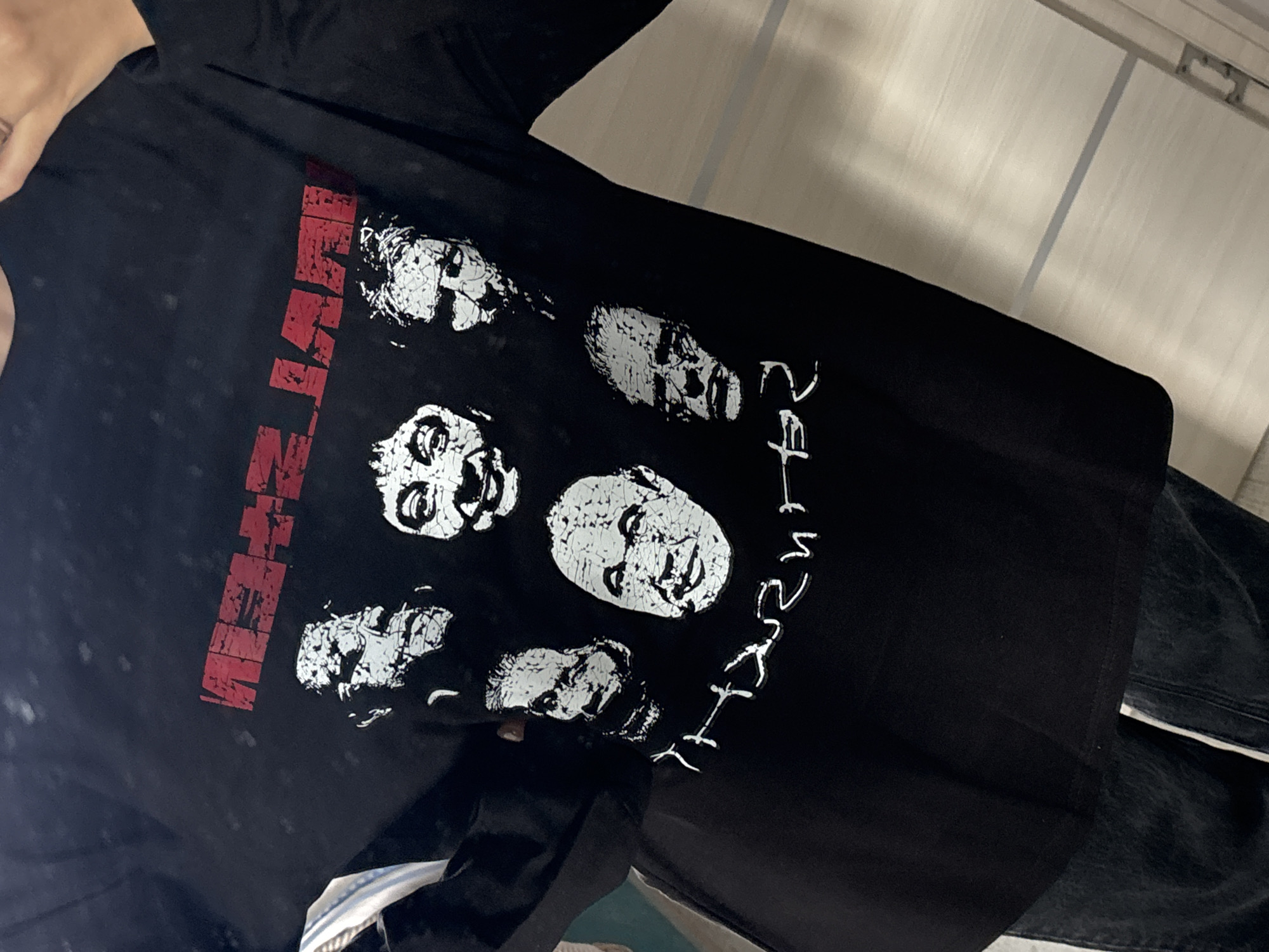 The image shows a black t-shirt with a graphic design printed on the front. The design includes several faces in a distressed, white ink style, and the name "MISFITS" printed in red. The t-shirt appears to be worn by someone. The environment includes clothing and a tiled surface, suggesting a domestic or retail setting. There is no evidence to suggest the picture was taken in Krasnoyarsk, Russia.