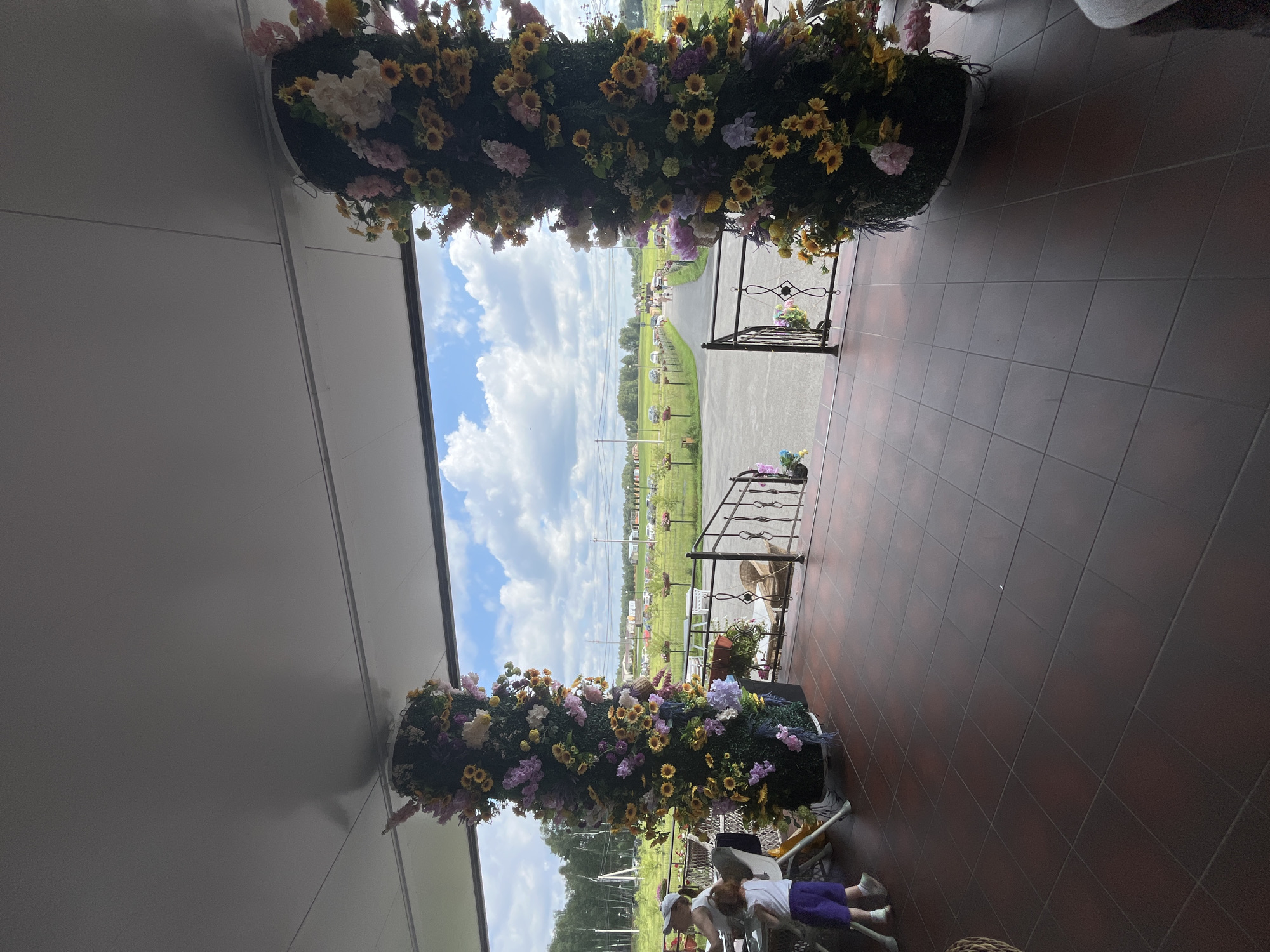 The image depicts an outdoor seating area with a tiled floor, likely at a restaurant or cafe, in Akhlebaikha (Kovriginskii S/S), Russia. Two large, cylindrical floral arrangements, adorned with sunflowers and other artificial flowers, stand prominently near the entrance to an outdoor pathway. In the foreground, a person wearing a white cap and a child in purple shorts and a white top are seated at a table. The pathway leads to a green field with scattered trees and a few parked cars visible in the distance under a partly cloudy sky. A black metal railing borders the steps to the pathway.