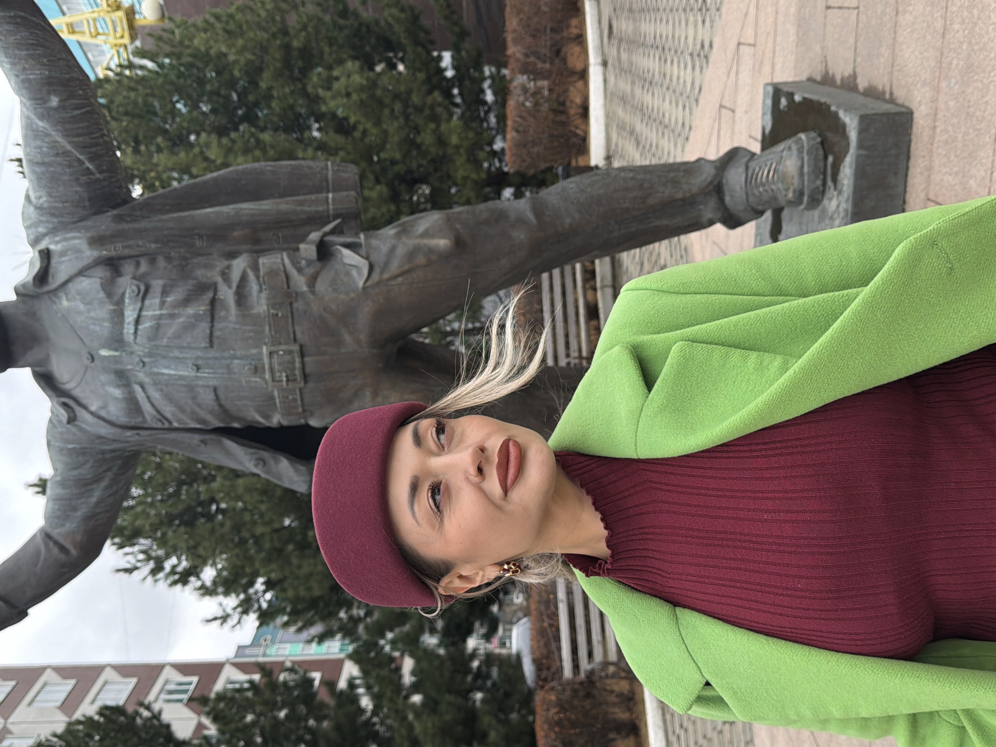The image depicts a woman standing near a bronze statue in what appears to be a public space. The woman is wearing a green coat over a maroon top and a matching maroon beret. She has fair skin and blonde hair, and is wearing gold earrings. She is looking to her left.
Behind her is a large bronze statue of a man in a coat, with his arms outstretched. The statue is set on a stone base, and there are trees and buildings in the background. The statue appears to be the "Uncle Yegor" statue on Prospekt Mira in Krasnoyarsk, Russia. There is pavement visible near the statue. The sky is overcast.