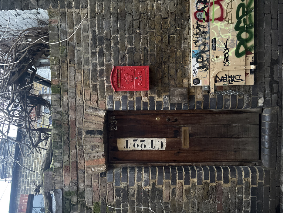 The image displays a detailed view of an old, weathered brick wall, predominantly composed of dark grey and brown bricks with occasional reddish hues and patches of moss.
On the far left, a dense tangle of bare, gnarled branches or vines is visible, suggesting a tree or shrub growing close to the building.
A vibrant red, ornate cast-iron mailbox is mounted on the brick wall, slightly to the left of the central axis of the image. It features decorative detailing, including what appears to be a crest or emblem in its center.
Below the red mailbox, a dark wooden door or panel is recessed within a semicircular brick archway. The door is constructed from horizontal wooden planks. On the left side of this wooden surface, the numbers "23A" are painted vertically in white. Centered across the wooden panel, there is a white rectangular plaque with the numbers "T92TD" painted in black, which appear upside down and reversed in the image. A brass-colored rectangular letter slot is also visible on the right side of the wooden panel.
To the right of the red mailbox, and extending down to the level of the wooden door, is a lighter-colored panel, possibly wood or metal, that is heavily covered in multi-colored graffiti tags and writings in black, green, red, blue, and pink. Several stickers are also affixed to this panel. A metal chain hangs from the bottom part of this graffiti-covered panel.
The scene appears to be illuminated by natural daylight. The combination of old brickwork, the bright red mailbox, the rustic wooden door, and the contemporary graffiti creates a contrast of textures and eras.