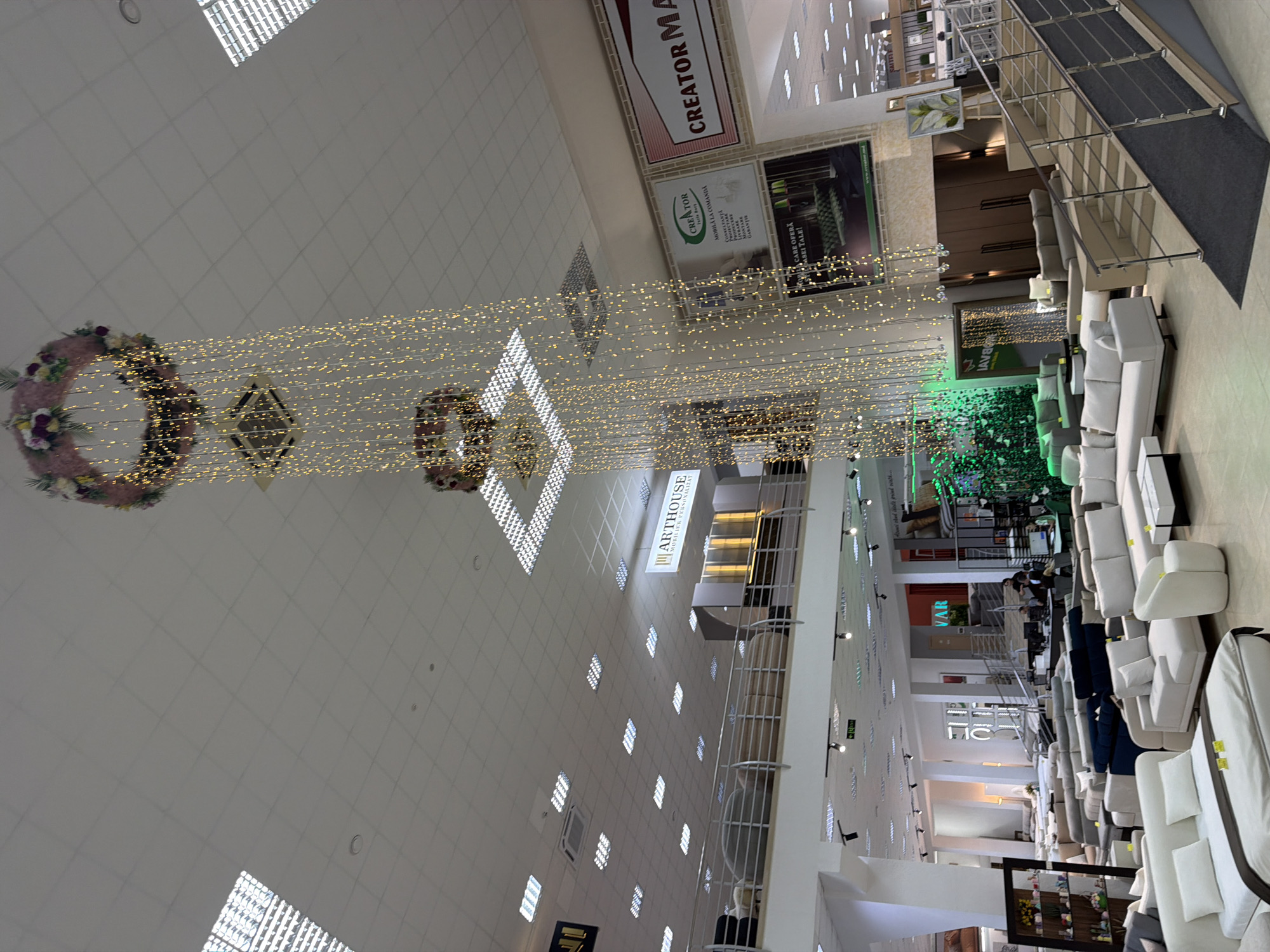 The image depicts the interior of a large furniture store, likely ARTHOUSE based on signage visible in the image, located in Trușeni, Moldova. The store features multiple levels, with a mezzanine level visible in the background. The foreground and midground are dominated by a wide array of sofas and seating arrangements of various styles and colors, including sectional sofas, modular sofas, and individual armchairs. A white sectional sofa with a bed configuration is prominently displayed in the left foreground.
The store's ceiling is a grid of fluorescent lights. Hanging from the ceiling are two large circular floral decorations with cascading strings of golden fairy lights, creating a festive atmosphere. In the midground, a large display unit with illuminated shelving is visible, along with various other furniture pieces like beds and cabinets.
Signage for "ARTHOUSE MOBILIER PERSONALIZAT" (ARTHOUSE PERSONALIZED FURNITURE) is present on the upper level. Other advertisements, such as for "CREATOR" and "CREATOR MA", are also visible.
A modern staircase with metal railings and a carpeted ramp leads up to another level on the right side of the image. A few people are sparsely distributed throughout the store, appearing to be customers or staff, some of whom are near the staircase and a sales counter in the background. A green living wall feature with integrated lighting is visible in the mid-background, adding a touch of nature to the commercial space. The overall impression is of a well-lit, spacious showroom showcasing a wide selection of home furnishings.