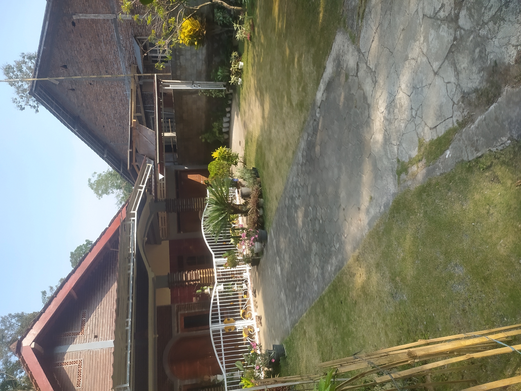 The image depicts a residential property located in Lopait, Indonesia. The scene features a pathway paved with hexagonal concrete stones leading from a white metal gate towards the entrance of a building. The pathway is bordered by a manicured lawn.
To the left of the pathway, there is a building with a reddish-brown brick facade and a dark red roof. The building has a covered porch area supported by pillars. A white metal gate with decorative elements stands in front of this building, adorned with potted plants, including a sago palm and flowering plants.
To the right of the pathway and beyond the gate, another building is visible, constructed with reddish-brown bricks and a dark roof. This structure appears to be a house or another part of the residential compound. The area to the right of the pathway is a well-maintained lawn with several potted plants and a larger tree with yellow foliage.
The sky is clear and blue, indicating a sunny day. The overall impression is that of a well-kept residential area, likely a private home in Lopait, Indonesia.