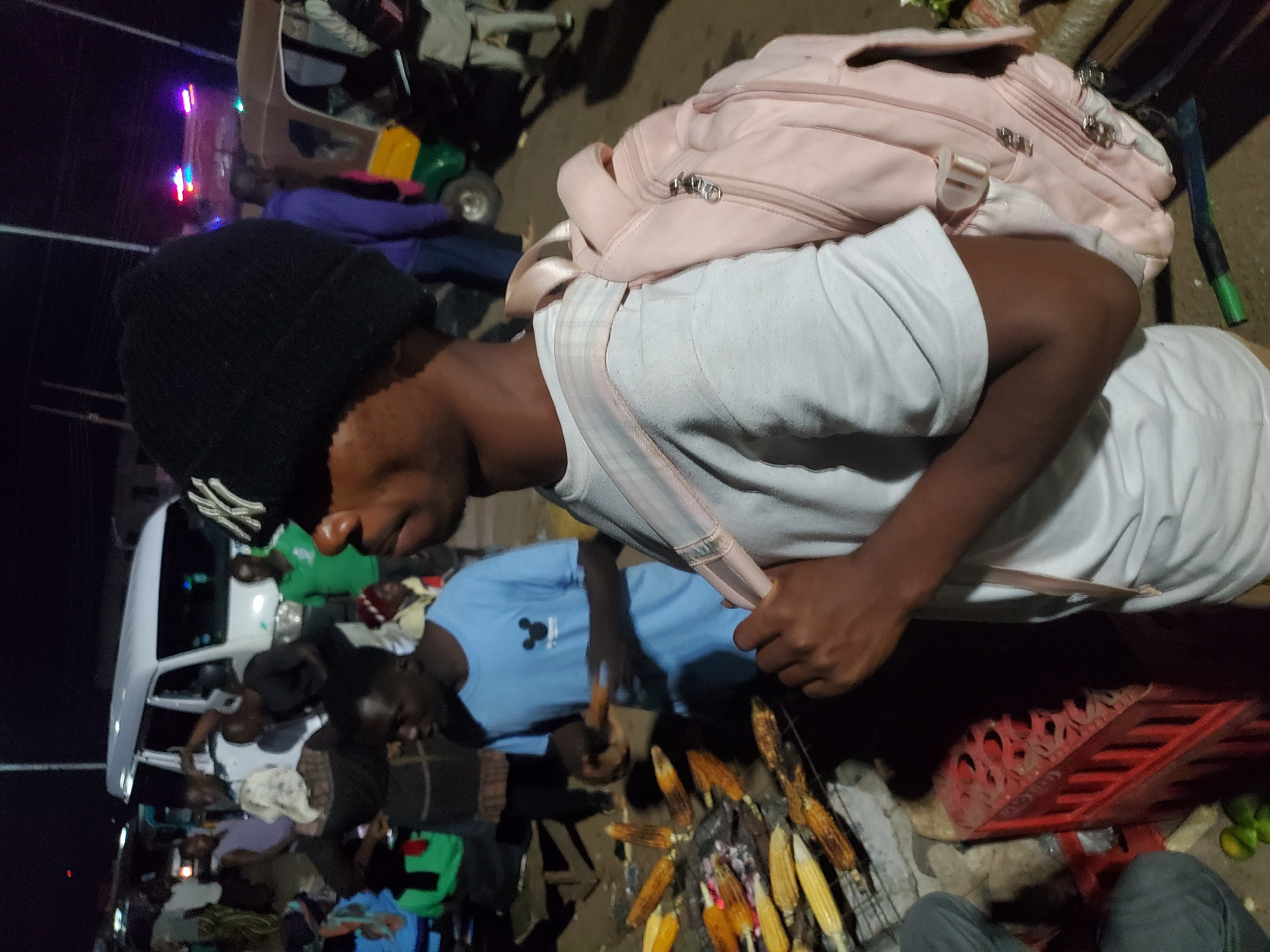 This night-time image, taken in Bukuru, Nigeria, captures a lively outdoor street scene, likely a market or food stall. The central figure, appearing to be a person with dark skin, wears a black beanie with a white "NY" (New York Yankees) logo and a white t-shirt. A light pink backpack with metallic zippers is slung over their right shoulder.
In the mid-ground, a significant amount of corn on the cob is seen roasting over hot coals on a grill, casting an orange glow. Another individual, wearing a light blue t-shirt with a black Mickey Mouse logo, is tending to the roasting corn. Numerous other people are visible in the background, some seated, some standing, contributing to the busy atmosphere.
The scene is dimly lit by artificial light sources, including colorful string lights (purple, blue, red) suspended overhead, and lights from vehicles in the distance. A white van or similar vehicle is partially visible on the left. On the ground to the right, a red plastic crate and several small green citrus fruits, possibly limes, are present. The overall impression is that of a bustling local street food vendor at night.