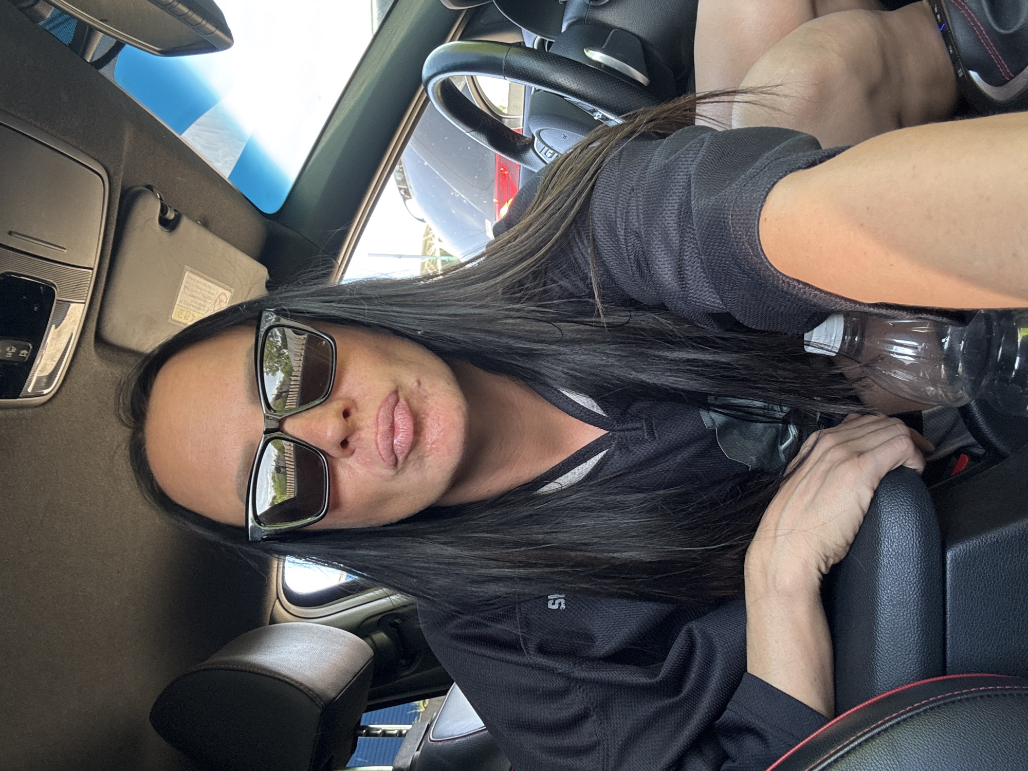 A woman with long dark hair, wearing sunglasses and a black athletic jersey, is sitting in the driver's seat of a car. She is holding a plastic water bottle in her right hand. The car's interior is visible, including the steering wheel, dashboard, and sun visor. The exterior is visible through the windows. There is no information in the image indicating that the location is Chaikovsky, Russia.