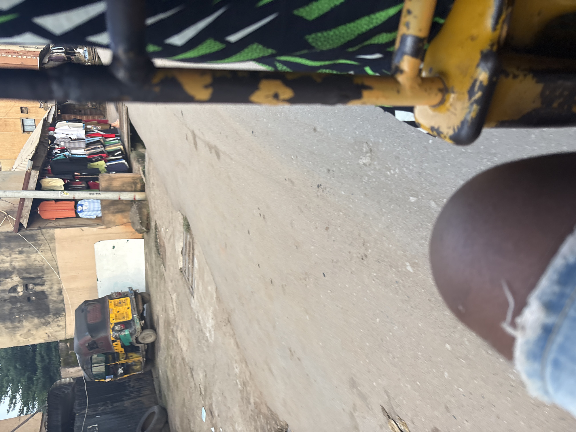 A first-person perspective shot from within a yellow and black autrickshaw in Onitsha, Nigeria, shows a street scene. The autrickshaw's yellow metal frame is visible in the foreground, with a person's knee and denim shorts on the lower left. To the left of the autrickshaw, a maroon and yellow autrickshaw is parked next to a wall. A market stalls with colorful clothing are displayed in front of a building on the right side of the street. The street itself is paved with asphalt and shows signs of wear and tear, with some debris scattered on the surface. The atmosphere appears to be typical of a bustling urban environment in Nigeria.