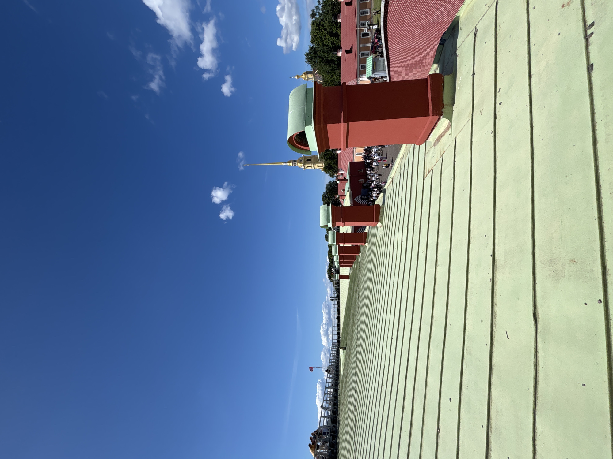 The image, taken from a high vantage point, depicts a bright, sunny day in Saint Petersburg, Russia. The sky is a vibrant, clear blue with scattered white clouds. In the foreground, a light green, corrugated metal roof stretches across the frame. The roof is punctuated by several red, rectangular structures, likely ventilation shafts or architectural elements, with rounded green tops.
Beyond the roof, a cityscape unfolds. A prominent feature is a golden spire, characteristic of Russian Orthodox churches, rising majestically against the blue sky. This spire is part of a larger complex of buildings with red roofs and some green-domed structures. Trees line the edge of the buildings, suggesting an urban park or a well-landscaped area.
Further in the distance, a railing or fence lines a walkway or viewing platform, with several people visible, suggesting a tourist area or public space. A red and white flag is flying from a pole on this platform. The overall scene suggests a popular tourist destination, offering panoramic views of the city's historical architecture. The time of day appears to be mid-morning to early afternoon, given the bright sunlight and clear skies. No text is visible in the image.