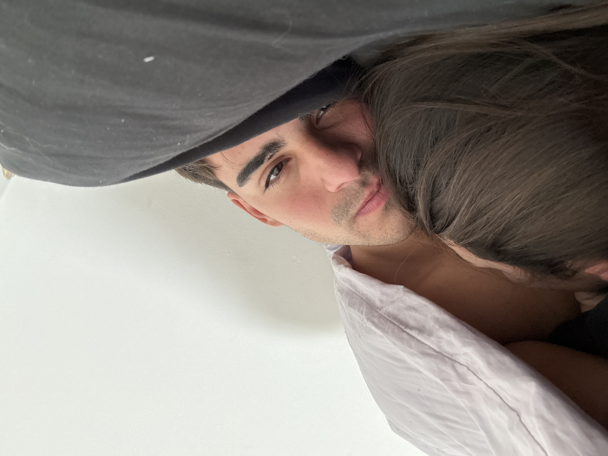 This is a close-up, overhead shot of a young person's face, their head resting on a light purple pillow. They are also partially covered by a dark gray piece of fabric, possibly a blanket or clothing. The person has dark brown hair and is looking directly at the camera with a neutral expression. Their eyes are brown and their eyebrows are well-defined. The lighting suggests it is daytime, and the setting appears to be indoors, given the white wall and pillow. The person is wearing a light-colored garment, possibly a shirt, with a slightly rumpled collar. The overall impression is intimate and personal, as if the photo was taken in a relaxed, private moment. There is no visible text in the image. The location context is Avellaneda, Argentina, but there are no specific visual cues within the image to confirm this.
