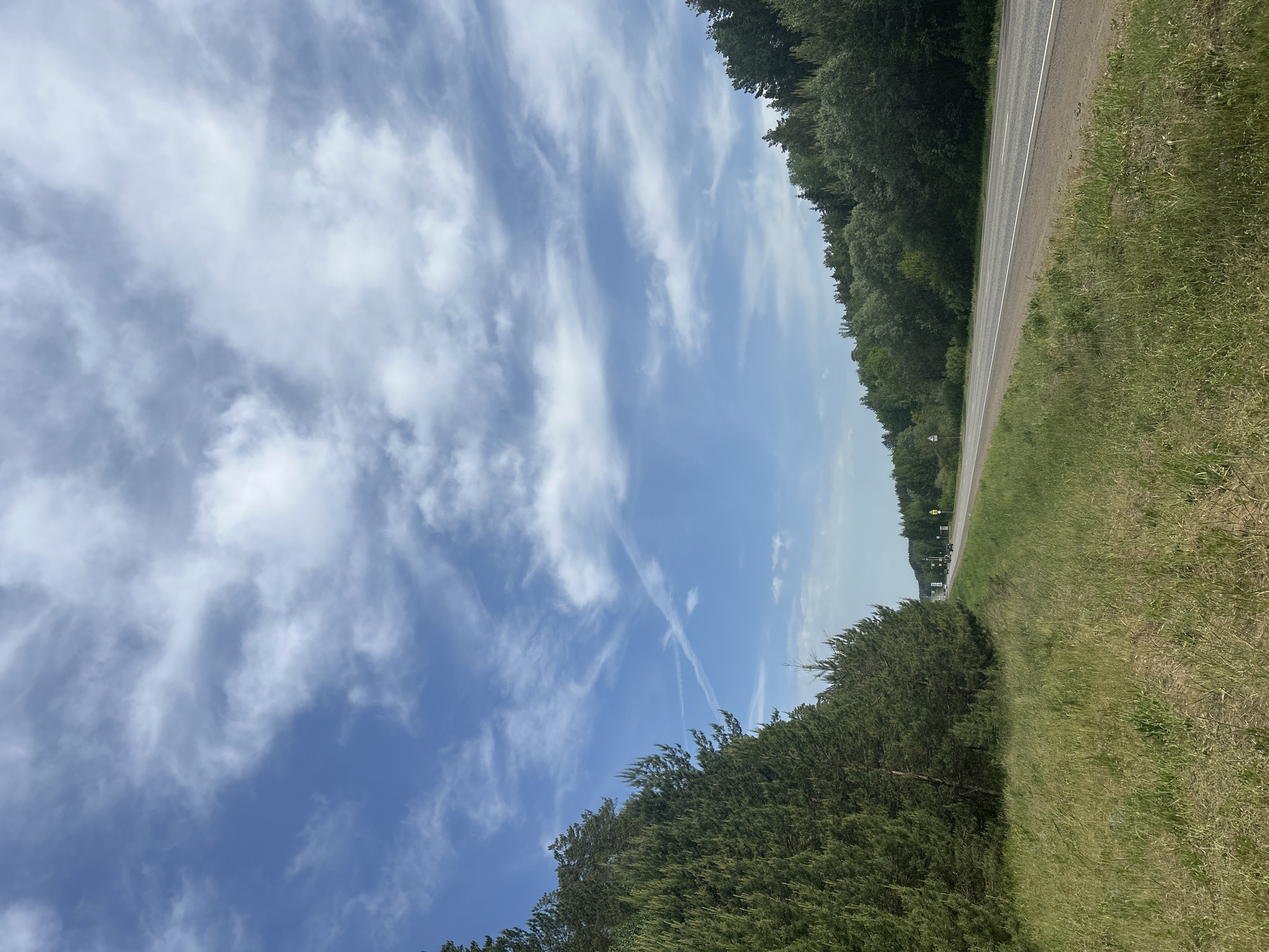 The image presents a landscape scene likely located near the city of Chaikovsky, Russia, based on the surrounding environment typical of the region. The sky occupies the major portion of the frame, featuring a vibrant blue hue streaked with white cirrus clouds and vapor trails from aircraft. A paved road runs along the right edge, bordered by dense green vegetation, possibly trees and thick grass. On the lower-left portion of the frame, there are trees. There are no people are visible in this shot.