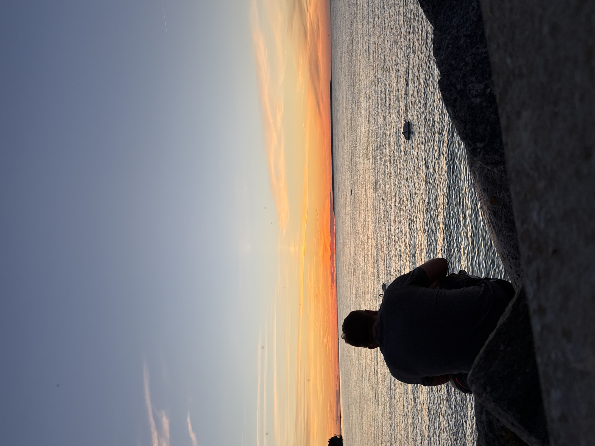 A man sits on rocks facing a calm sea at sunset. The sky is a gradient of blues and oranges, with thin clouds. In the distance, a dark landmass is visible on the horizon. A small boat is in the water to the right of the man. The foreground is dominated by dark rocks. The image does not contain any identifiable landmarks of Tallinn, Estonia.