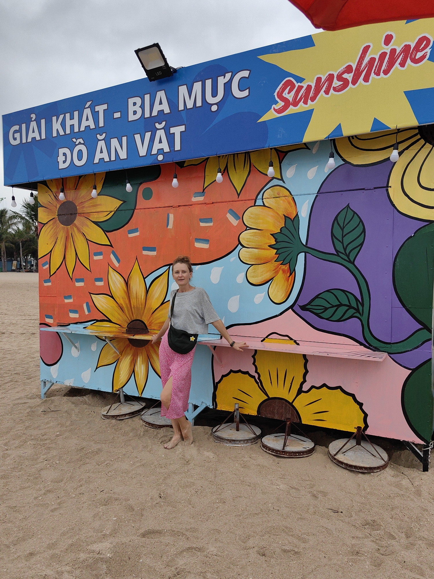 A woman stands barefoot on a sandy beach, posing casually in front of a brightly painted stall. The stall is adorned with a vibrant mural featuring large, stylized sunflowers and other floral patterns in shades of yellow, orange, purple, green, and blue, along with abstract shapes like raindrops and rectangles.
A blue signboard runs along the top of the stall, displaying white text in Vietnamese: "GIẢI KHÁT - BIA MỰC" (Refreshments - Squid Beer) and "ĐỒ ĂN VẶT" (Snacks). To the right, a yellow starburst shape holds the word "Sunshine" in red script. A floodlight, marked "LED," is mounted on the signboard, and several white light bulbs hang beneath it, along the stall's edge.
The woman is dressed in a grey t-shirt, a pink patterned skirt, and a black fanny pack, leaning against a small, pink counter extension of the stall. She has a neutral, relaxed expression. The ground is soft, light brown sand. In front of the stall, three rusty, round metal bases are partially buried in the sand, possibly serving as supports for tables or seating. Palm trees are visible in the background on the left, under an overcast sky, suggesting a cloudy daytime scene. The setting is a beach, specifically in Bai Chay Ward, Vietnam.