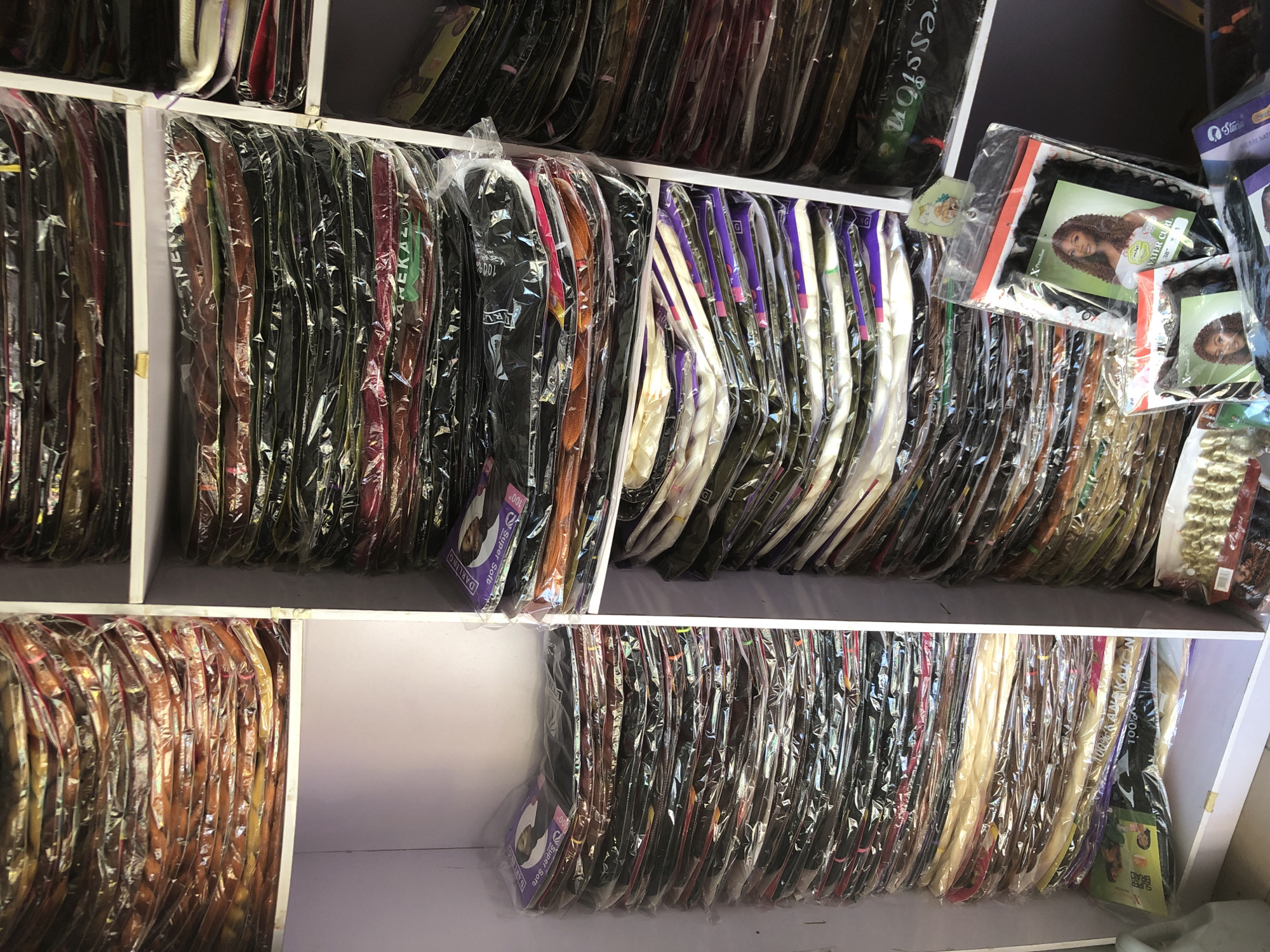 The image depicts a retail store in Ado, Nigeria, specializing in hair extensions. The store features shelves stacked high with numerous packages of braided and woven hair extensions. The products are predominantly packaged in clear plastic, revealing a variety of colors, including black, brown, blonde, orange, and purple. Some packages have labels with brand names and images of hairstyles, such as "DARLING SUPER SOFT" and "100% KANEKALON". In the foreground, a few individual packages of hair extensions are displayed, one featuring a model with curly dark hair and the label "WATER CURL". The overall impression is of a well-stocked establishment catering to the beauty and hair care needs of its customers.