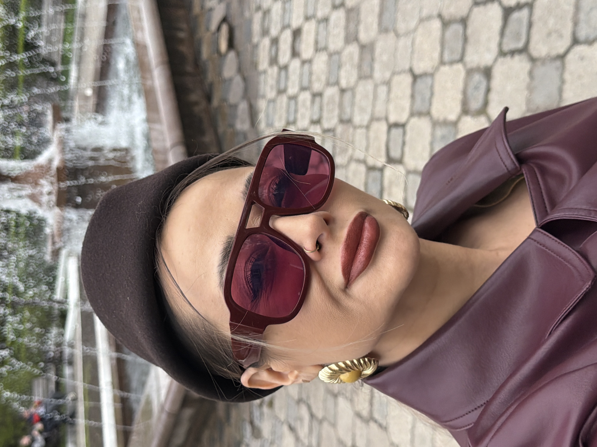 A woman is pictured outdoors, wearing a brown beret, sunglasses with burgundy frames, and a burgundy leather-like jacket. She has light skin and is wearing gold, flower-shaped earrings. A waterfall is visible in the background to the left. The background to the right appears to be a paved area with light grey rectangular stones. The photograph was taken in Krasnoyarsk, Russia.