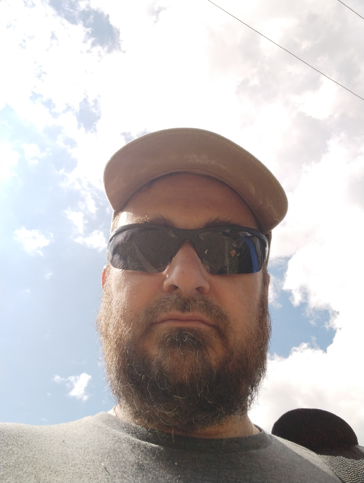A man is taking a selfie outdoors. He is wearing a tan cap and sunglasses, and has a beard. The background is a bright sky with white clouds, and there are power lines visible. The lower part of the image shows the man's grey shirt and a dark object, possibly another cap, on his shoulder. The image does not contain information about the specific location within Ширяево (ильинский), Russia.