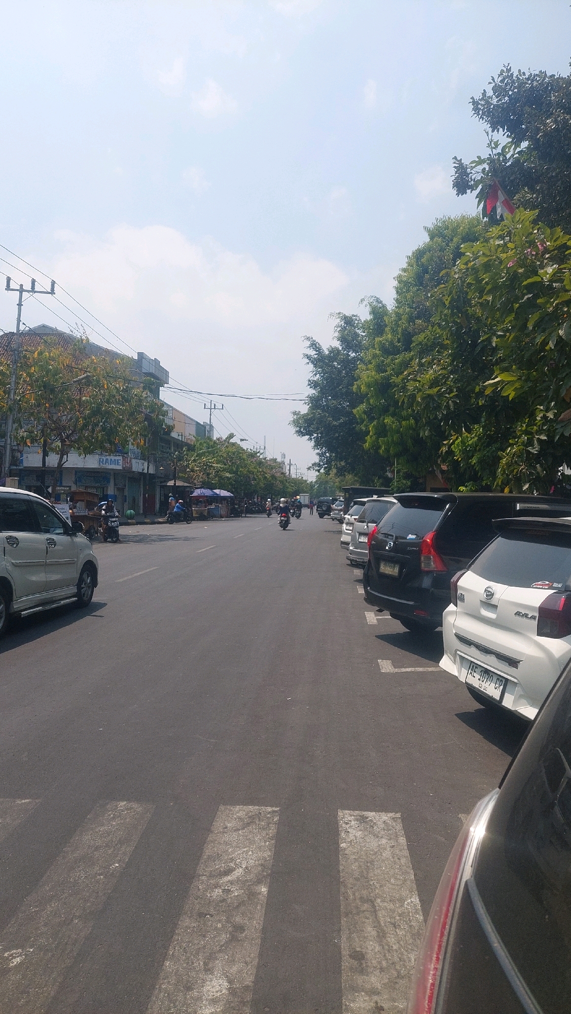 The image depicts a street scene in Madiun, Indonesia, likely a commercial area. The foreground shows a pedestrian crossing with worn white stripes on asphalt. To the right, several cars are parked parallel to the street, including a white car with a license plate reading "AE 1079 CR". Further down the street, a line of parked cars continues. On the left side of the street, a white van is visible, and beyond it, buildings with storefronts line the road. The buildings have awnings and signs, with one sign partially visible reading "RAME". Trees with lush green foliage are present along both sides of the street, casting shadows on the road. Several motorcycles are being ridden on the street, and a few people are visible walking or standing near the storefronts and stalls. The sky is bright blue with scattered white clouds, indicating it is daytime and likely sunny. The overall impression is of a moderately busy urban street with a mix of vehicles, pedestrians, and commercial establishments.