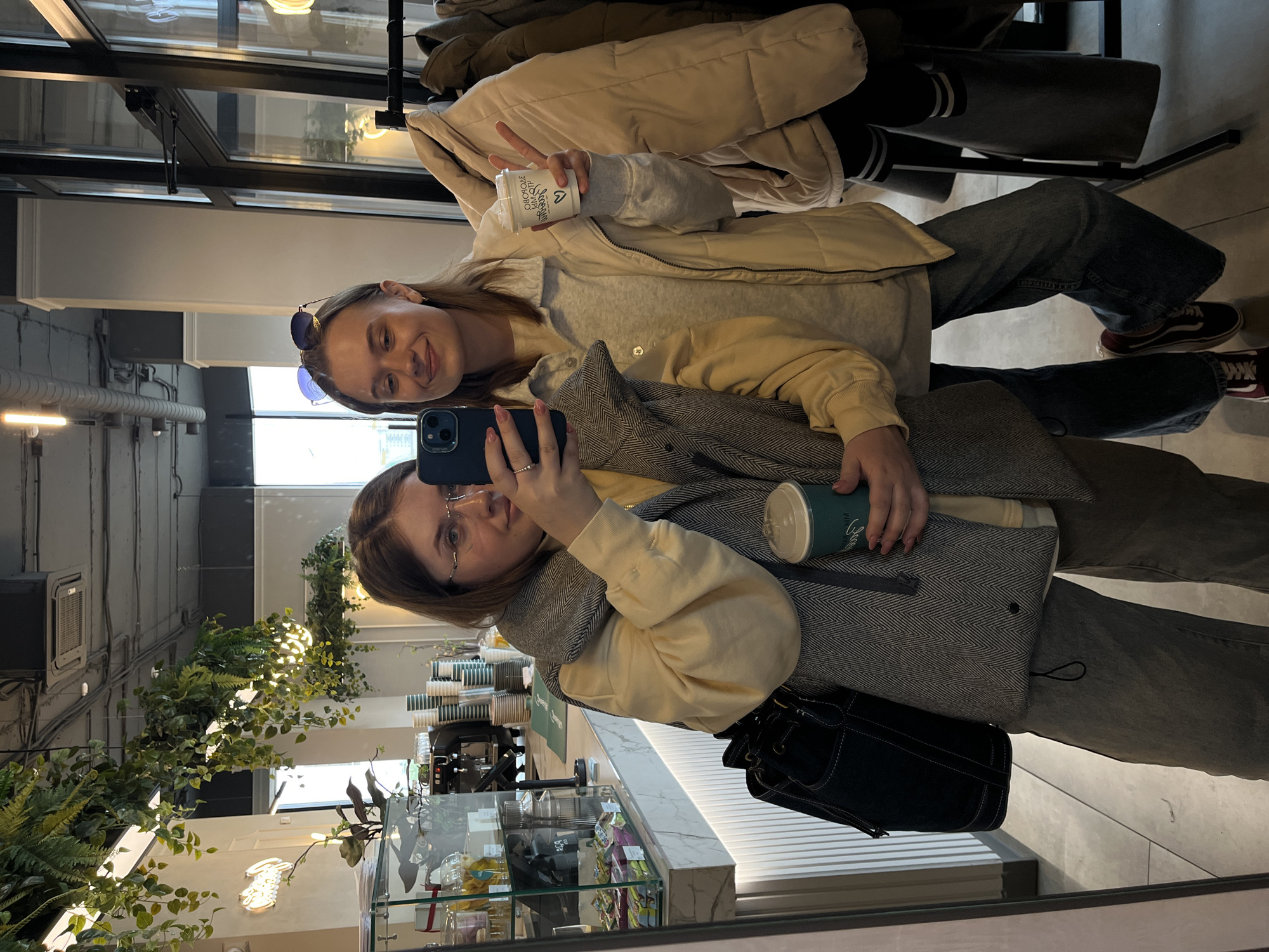 Two women are taking a selfie in a cafe. The woman in the foreground is holding a blue phone and wearing glasses, a light yellow sweatshirt, and a herringbone jacket. A black purse is near her. She's also holding a green cup. The other woman is wearing sunglasses on her head, a light yellow sweatshirt, and a beige puffy jacket. She's holding a white cup and making a peace sign. They are both wearing jeans and sitting near a rack of clothes. A marble countertop is visible with an espresso machine and refrigerated display case. The cafe is decorated with plants. The location is Krasnoyarsk, Russia.