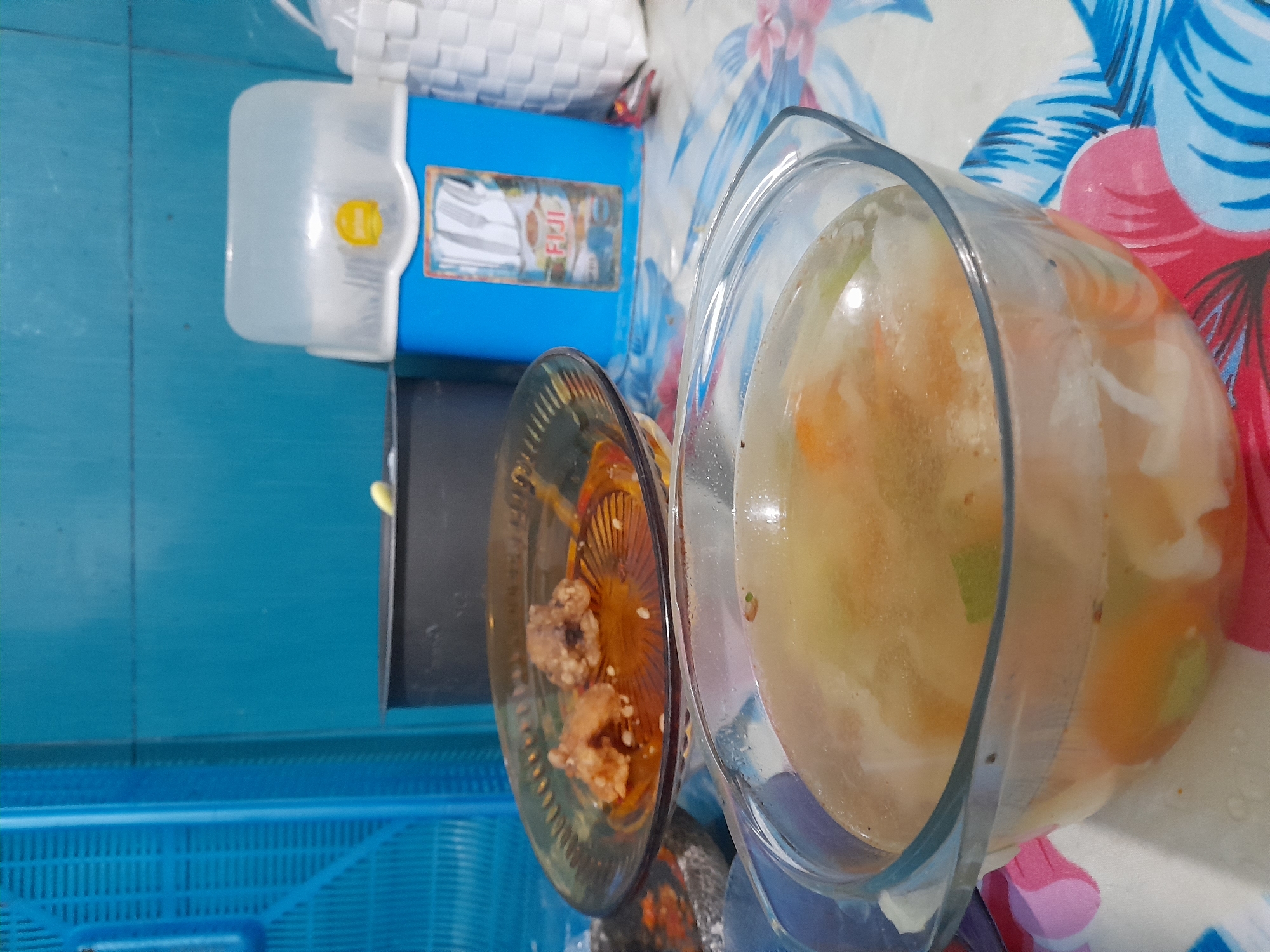 A top-down, rotated view depicts a domestic scene, possibly indoors, in Gelam, Indonesia. A glass casserole dish filled with a yellowish broth containing visible vegetables rests on a surface covered with a floral-patterned cloth. Adjacent to the dish is a brown glass plate holding pieces of what appears to be fried food, possibly chicken. To the left of the plates are a blue plastic cutlery holder with a white top, and a black cutting board. A blue plastic basket is partially visible in the bottom left corner. The background includes a blue surface, possibly a wall or table.