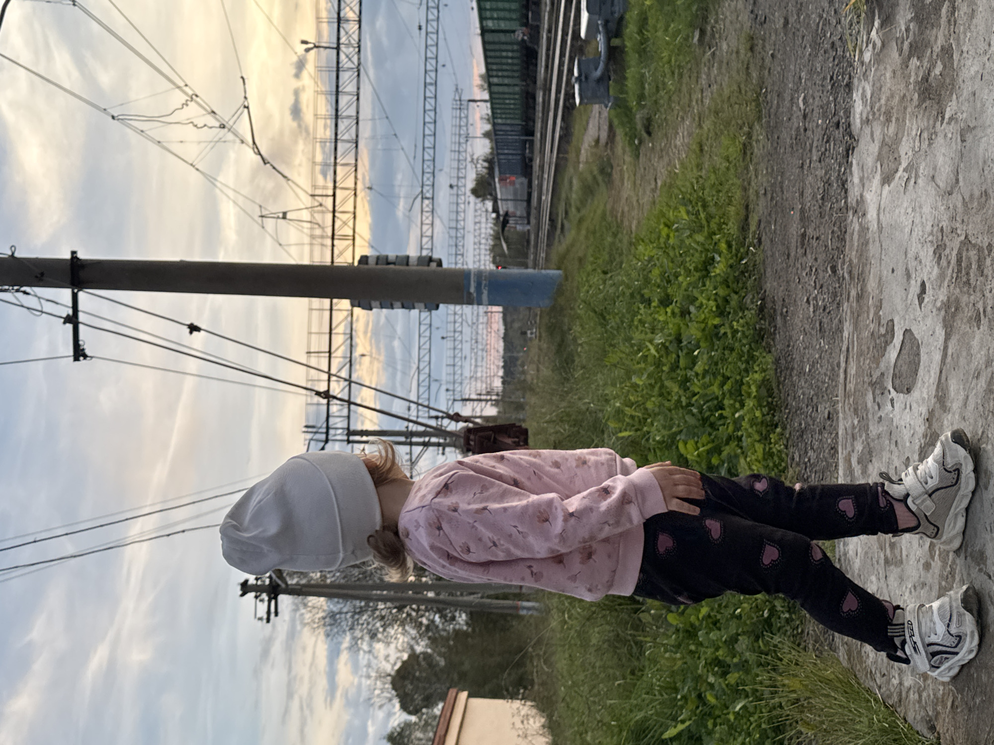 A young child, viewed from the side and slightly from behind, stands on a concrete platform. The child wears a white beanie, a pink sweatshirt with a floral pattern, black leggings with pink heart prints, and white and gray sneakers. The child faces a railway, with tracks and overhead electrical wires visible. A train is present on the tracks in the distance, consisting of multiple green cargo cars. The environment appears to be a rural or semi-rural area, possibly a train station, with overgrown grass and vegetation surrounding the tracks. The sky is overcast with hints of sunset colors peeking through the clouds. A building is partially visible to the left of the frame. The scene is captured outdoors during daylight.