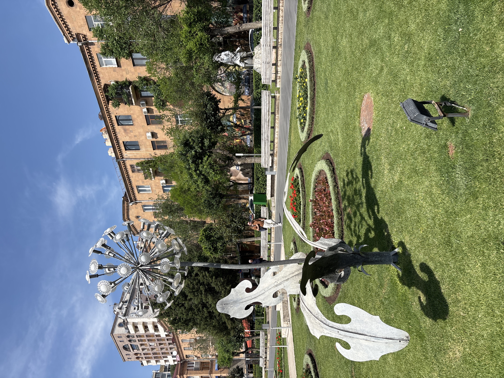 The image shows a sunny day in a park in Yerevan, Armenia. In the foreground, there is a large metallic sculpture of a dandelion, with a tall stem and a head made of many small glass-like structures. To the right, a man in a light brown shirt and white pants is walking on a paved path. In the background, there are several apartment buildings with orange-brown facades and trees. There is also a statue of a person in military attire standing on a pedestal, and several park benches. The park has well-maintained green lawns and flower beds. The sky is blue with scattered white clouds. The overall scene depicts a pleasant urban park environment.