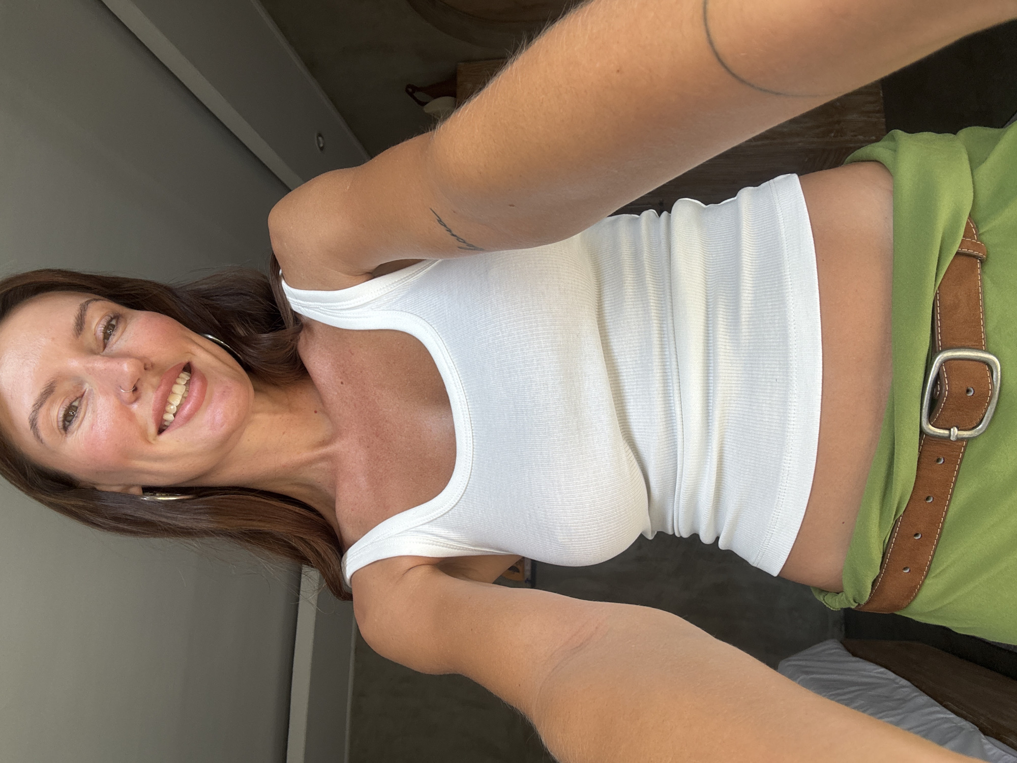 In North Kuta, Indonesia, a woman with long brown hair and a fair complexion is pictured from the chest up, facing the camera directly. She is wearing a white ribbed tank top and a green skirt cinched with a brown belt. A silver hoop earring is visible in her left ear, and a nose ring is in her right nostril. Her gaze is directed at the camera, and she has a bright smile, revealing her teeth. The background appears to be an indoor setting, with soft lighting and a hint of a window or doorway to her left, casting shadows on the wall.