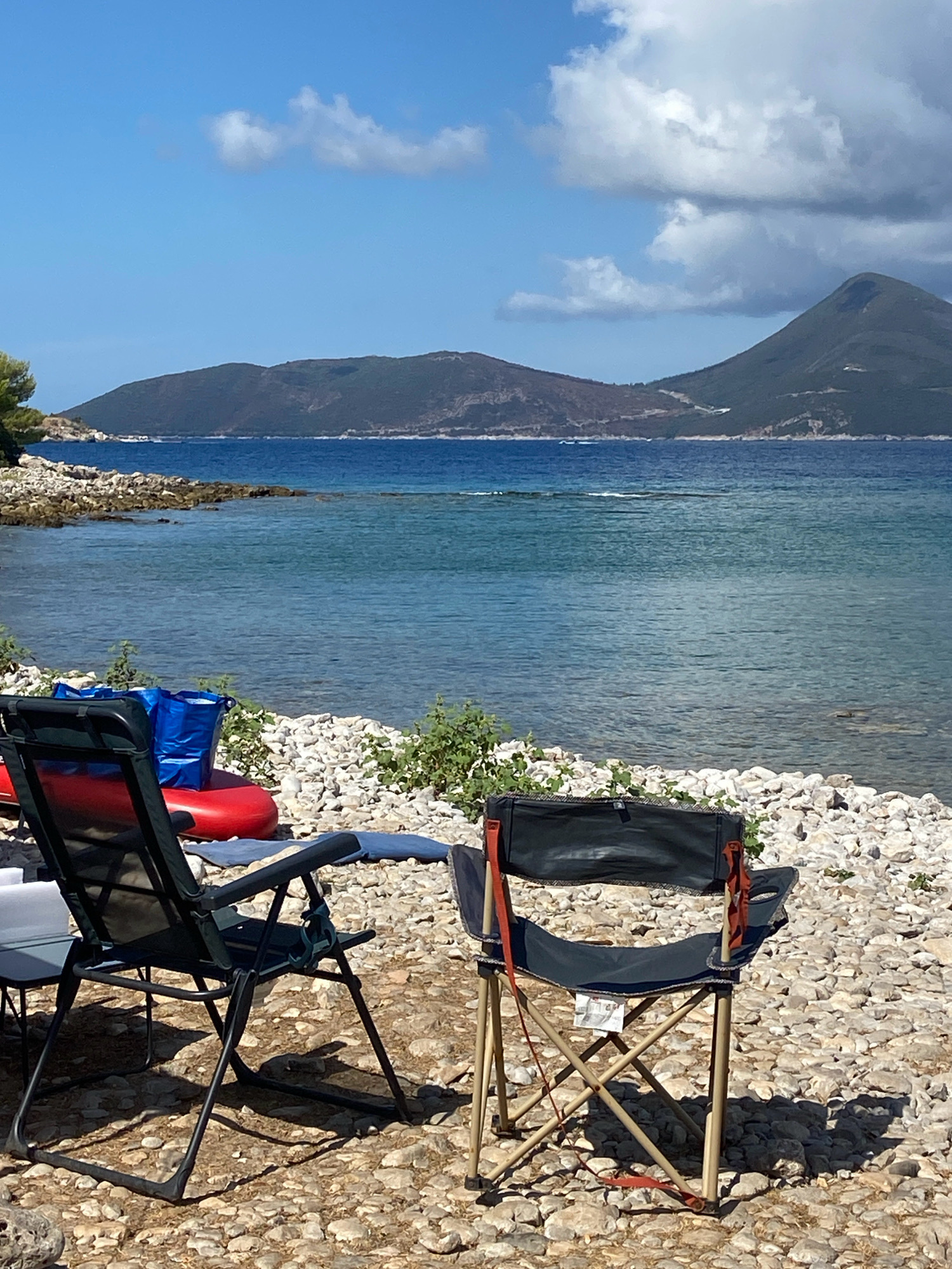 This is a vertical, eye-level shot of a rocky beach on the Luštica peninsula in Montenegro. The scene is set with two folding chairs in the foreground, one dark green and one dark gray, facing a tranquil, blue sea. Behind the chairs, a red inflatable raft and two blue bags are visible. The beach is covered in light-colored pebbles, with sparse green vegetation growing amongst them. In the mid-ground, the clear blue water stretches out, meeting the distant shoreline of a rugged, green mountain range under a bright blue sky dotted with fluffy white clouds. The overall impression is one of a peaceful, outdoor recreational setting by the sea.