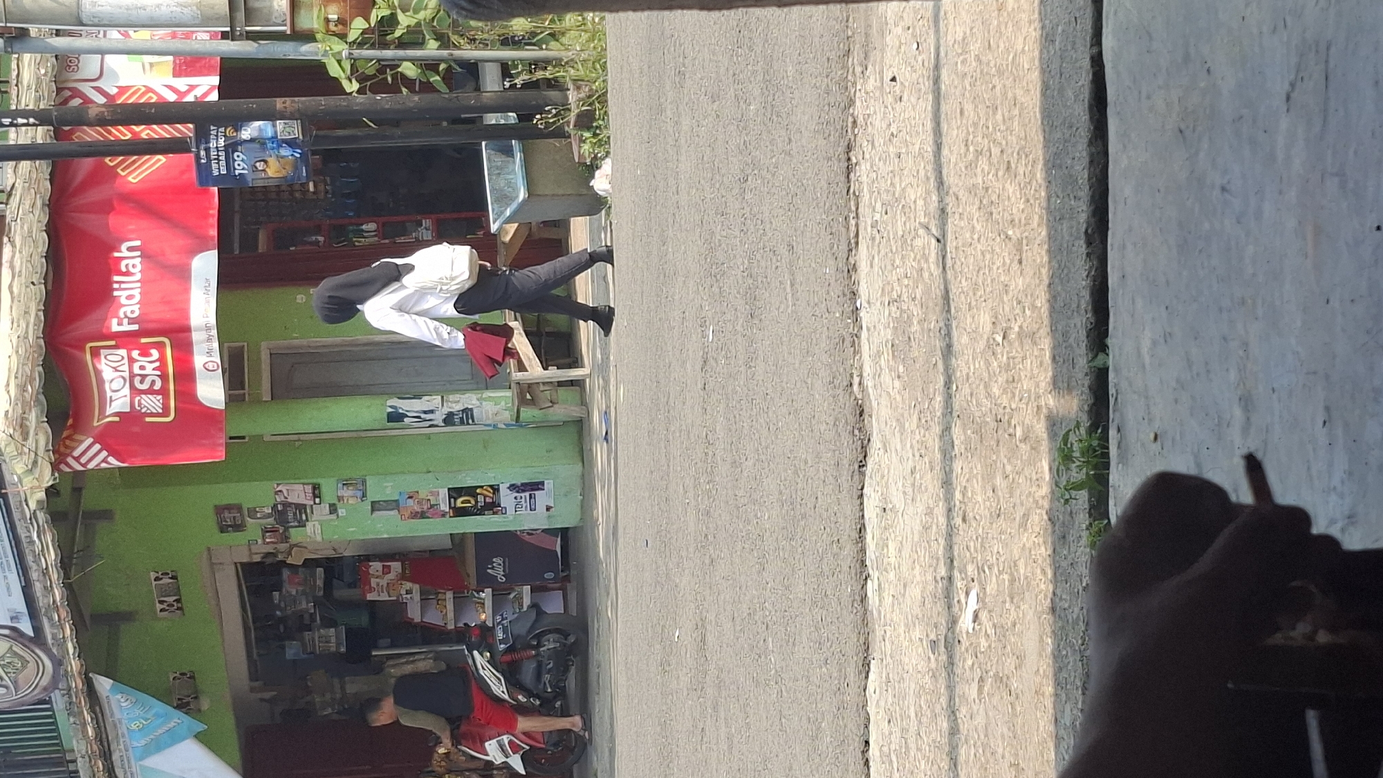 A street-level, eye-level, outdoor, daylight, long shot captures a scene on a street in Cilaku, Indonesia. In the background, a vibrant green building with a red banner featuring the text "TOKO SRC Fadilah" is visible. A person wearing a black hijab, white long-sleeved shirt, dark pants, and carrying a white backpack walks away from the building towards the right of the frame. They are also holding a red garment. In front of the building, a man in red shorts and a dark t-shirt is seated on a motorcycle. The street in the foreground is asphalt, and a concrete curb separates it from the background. The lighting suggests it is a sunny day.