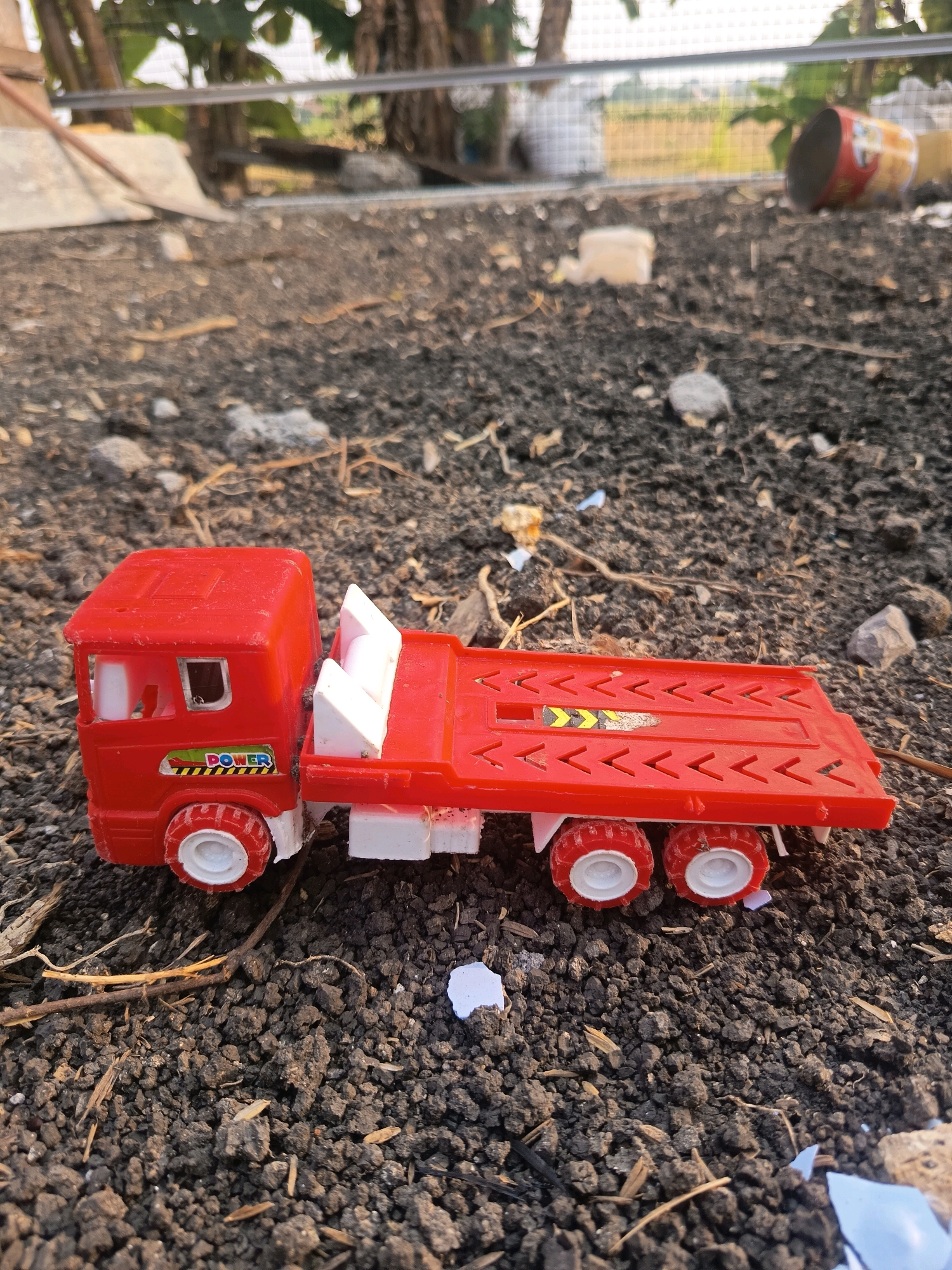 A red toy flatbed truck sits on dark, rough soil. The truck has a red cab with a white interior and a white trailer. The trailer is red with a textured surface. The truck has white wheels with red rims. The scene appears to be outdoors, possibly in an undeveloped or construction area, with dried leaves and small rocks scattered on the ground. In the background, out of focus, there are some green plants and what looks like a fence or structure. The lighting suggests it is daytime.