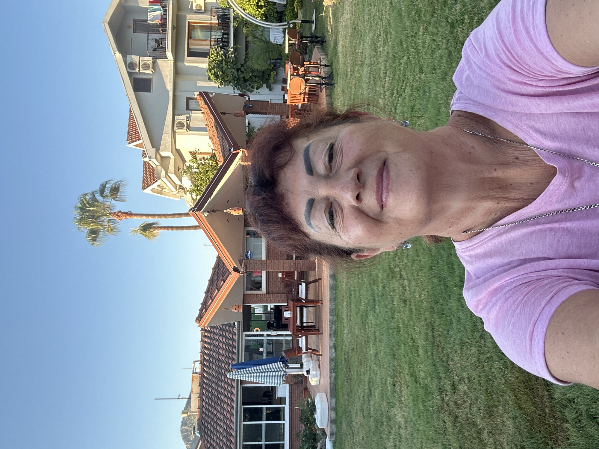 A mid-shot selfie shows a smiling woman with auburn hair and fair skin in the foreground. She is wearing a pink t-shirt and a silver necklace. Her eyes are closed, and she has prominent eyebrows. Behind her, a green lawn stretches out, leading to a patio area with wooden tables and chairs. Beyond the patio, there are buildings with red-tiled roofs and white walls, some with visible air conditioning units. Two palm trees are visible in the background. The scene appears to be outdoors on a sunny day. The location is described as Ortaca, Turkey, suggesting a warm, possibly coastal environment with Mediterranean architecture.