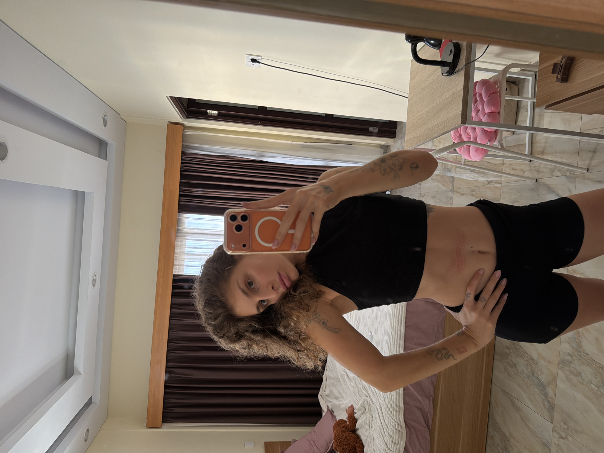 A mirror selfie captures a young woman posing in what appears to be a modern bedroom or studio apartment. The woman has curly brown hair and wears a black crop top and black shorts, revealing her midriff and tattoos on her arms—a floral design on her upper left arm, a snake on her lower right arm, and a small butterfly on her right hand. She is holding a peach-colored smartphone with a prominent white stylized letter "C" on its case, using it to take the photo.
The background shows a wooden bed frame with a light purple sheet and a white textured blanket, on which a small brown teddy bear rests. To her right, a wooden desk is visible with a black lamp, and a white metal chair with a pink, puffy cushion is positioned underneath. The room features white walls, a white ceiling with recessed lighting, and a light beige tiled floor with a marble-like pattern. Dark brown curtains or blinds cover a window. A wall socket with a black cable plugged in is seen on the wall above the window. The overall lighting is bright and even, suggesting daytime. The woman's expression is neutral, characteristic of a posed selfie. A small tattoo resembling "B3" is visible on her right forearm. The scene aligns with contemporary interiors often found in places like North Kuta, Indonesia, as specified by the context.